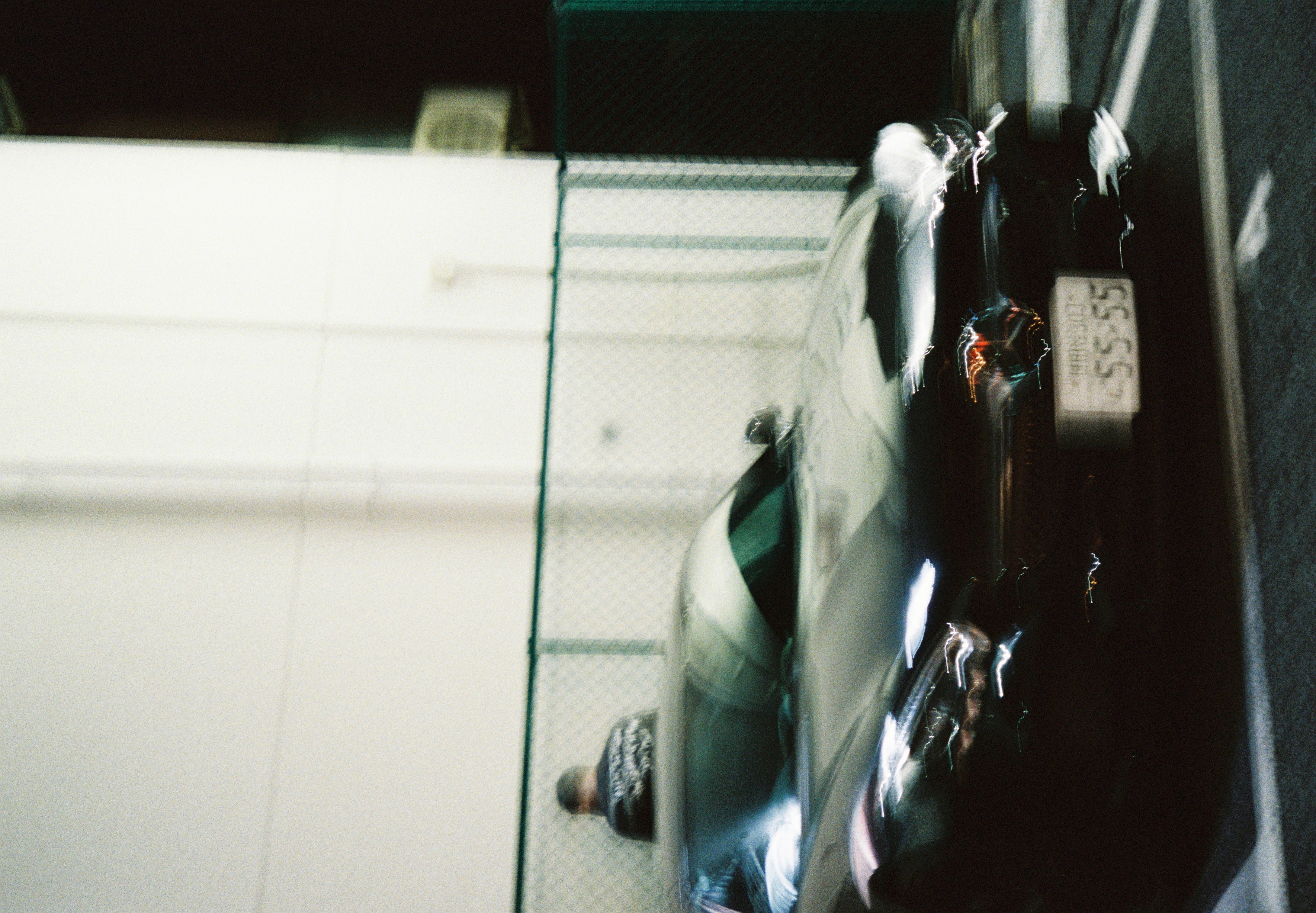 A dark, low sports car with a visible '55-55' license plate passes a chain-link fence, motion blur streaking its lights.