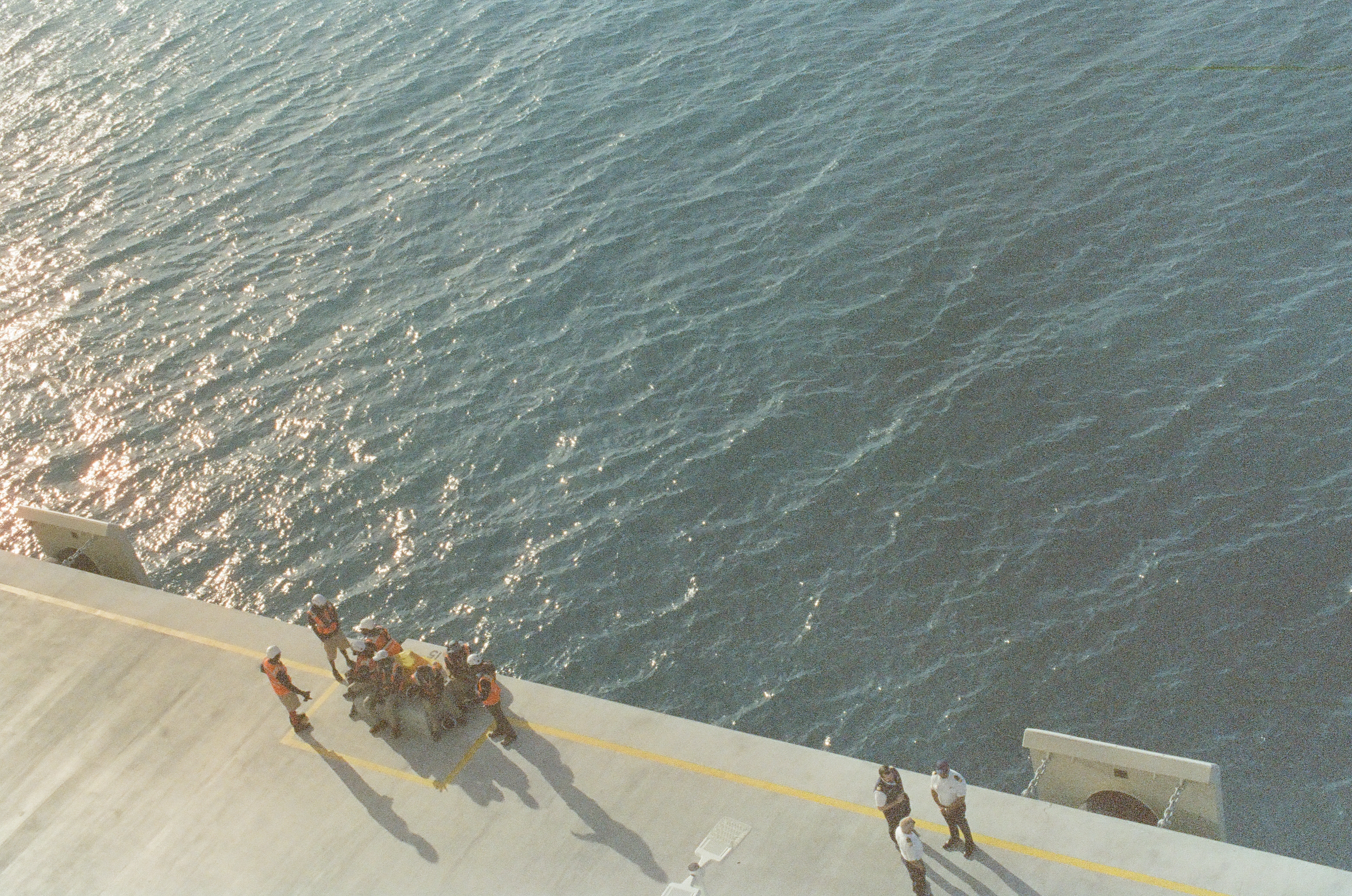Seen from above, workers in orange vests cluster near the edge of a pale paved surface beside dark rippling water, casting long shadows.