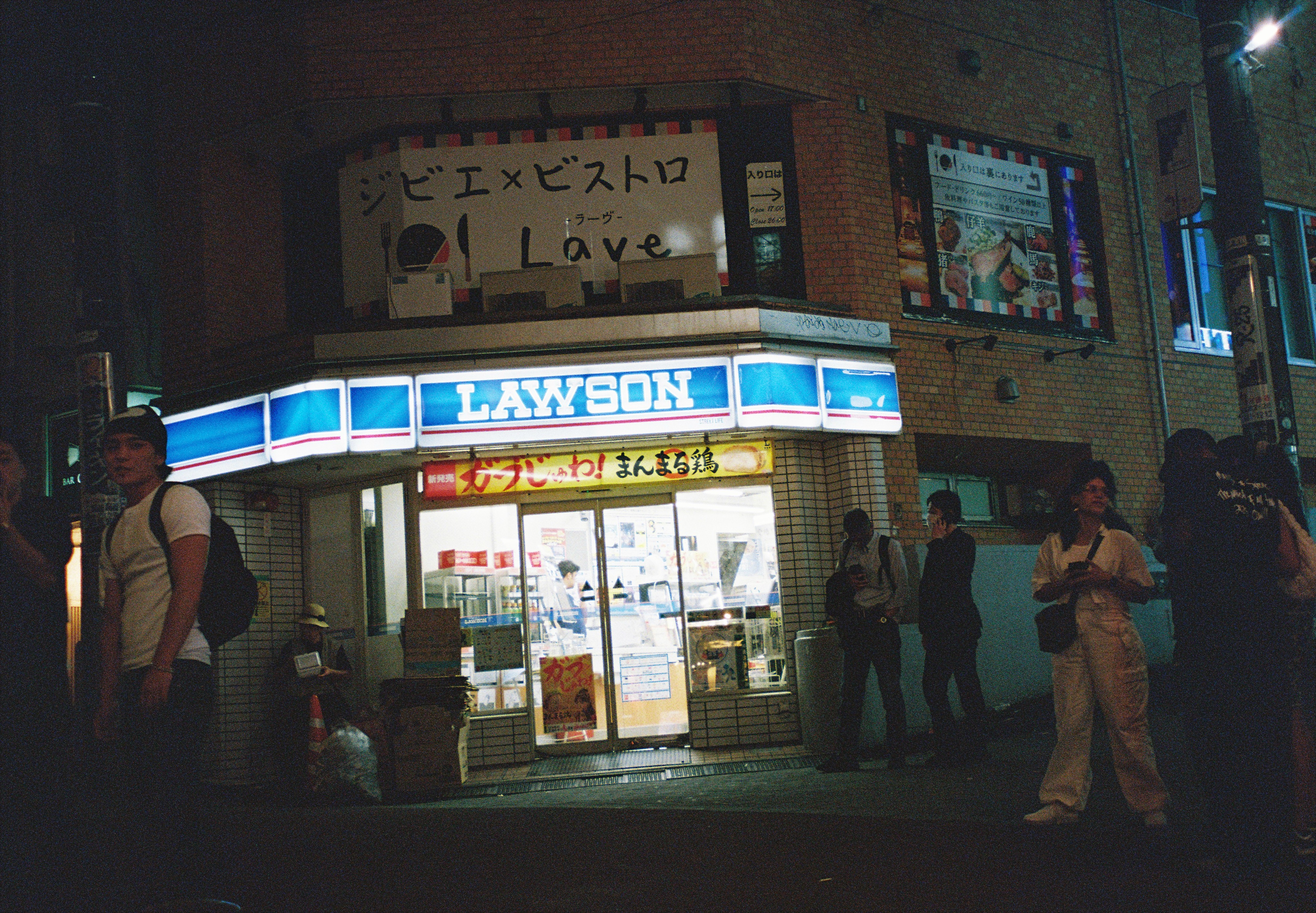 A brightly lit Lawson convenience store sign wraps around a corner storefront at night, with a hand-painted banner above reading 'Lave'.