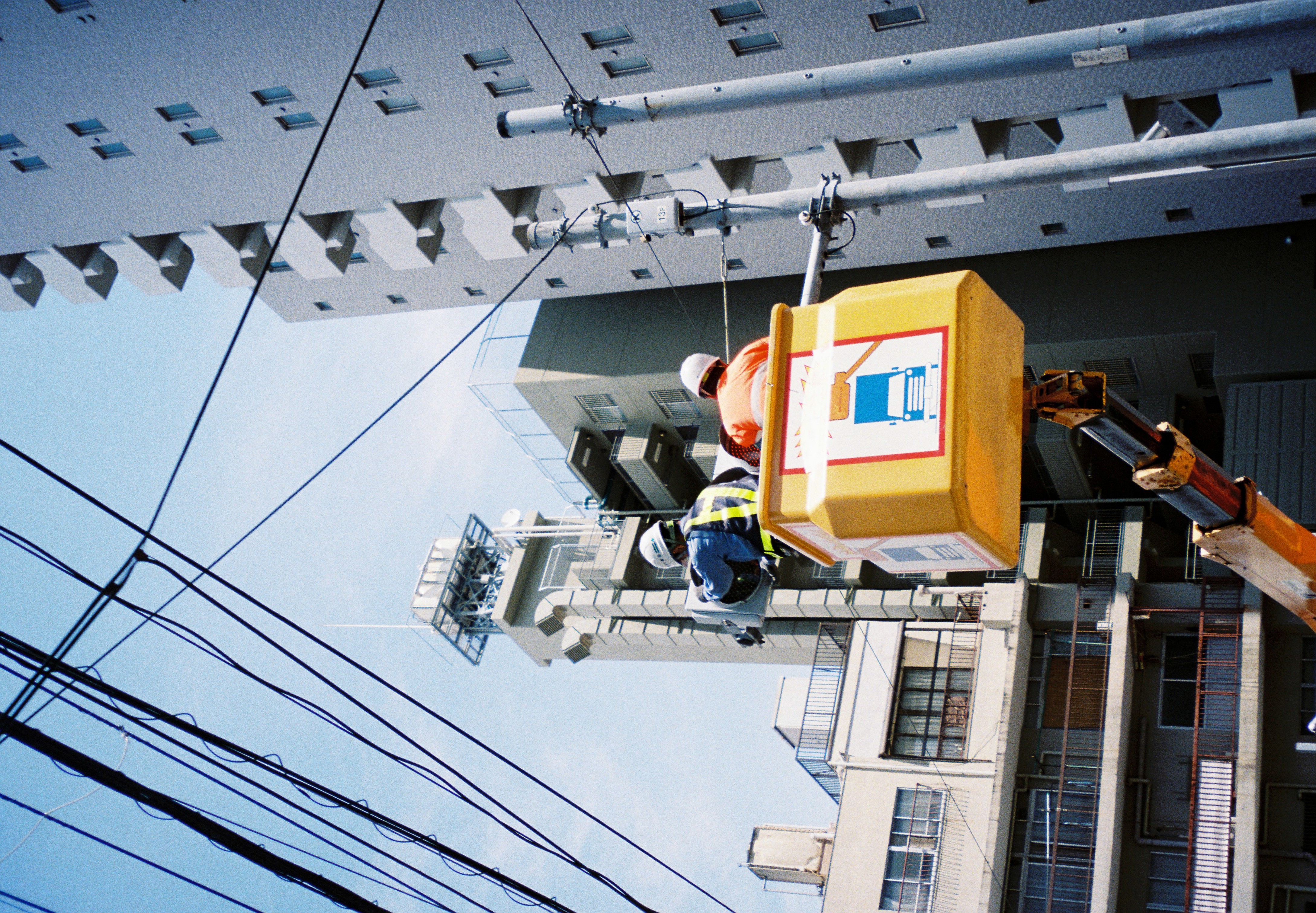 Two helmeted workers ride a yellow bucket lift up through a dense web of overhead wires beside tall apartment buildings.