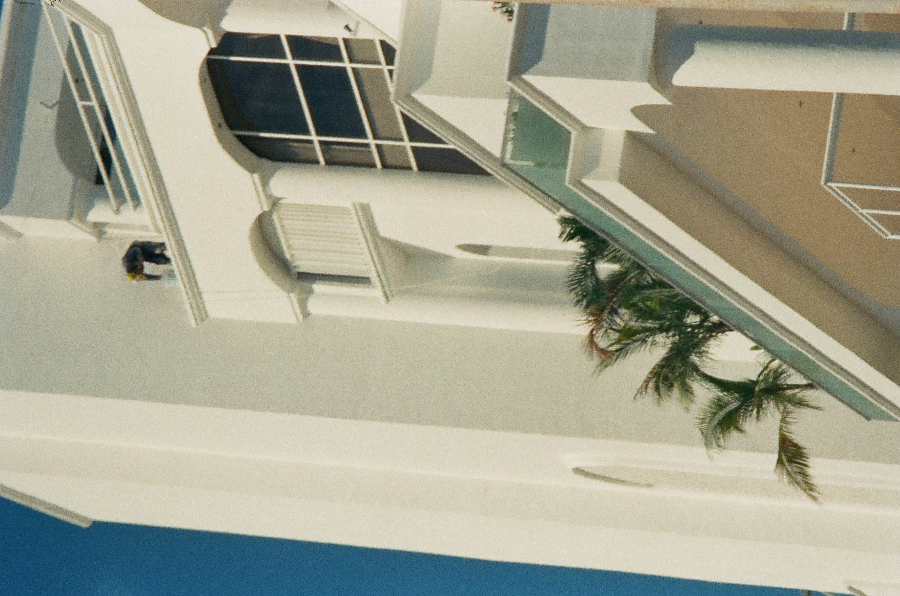 A small crouched figure appears near an upper ledge on a tall white building with arches, balconies, columns, windows, and palms.