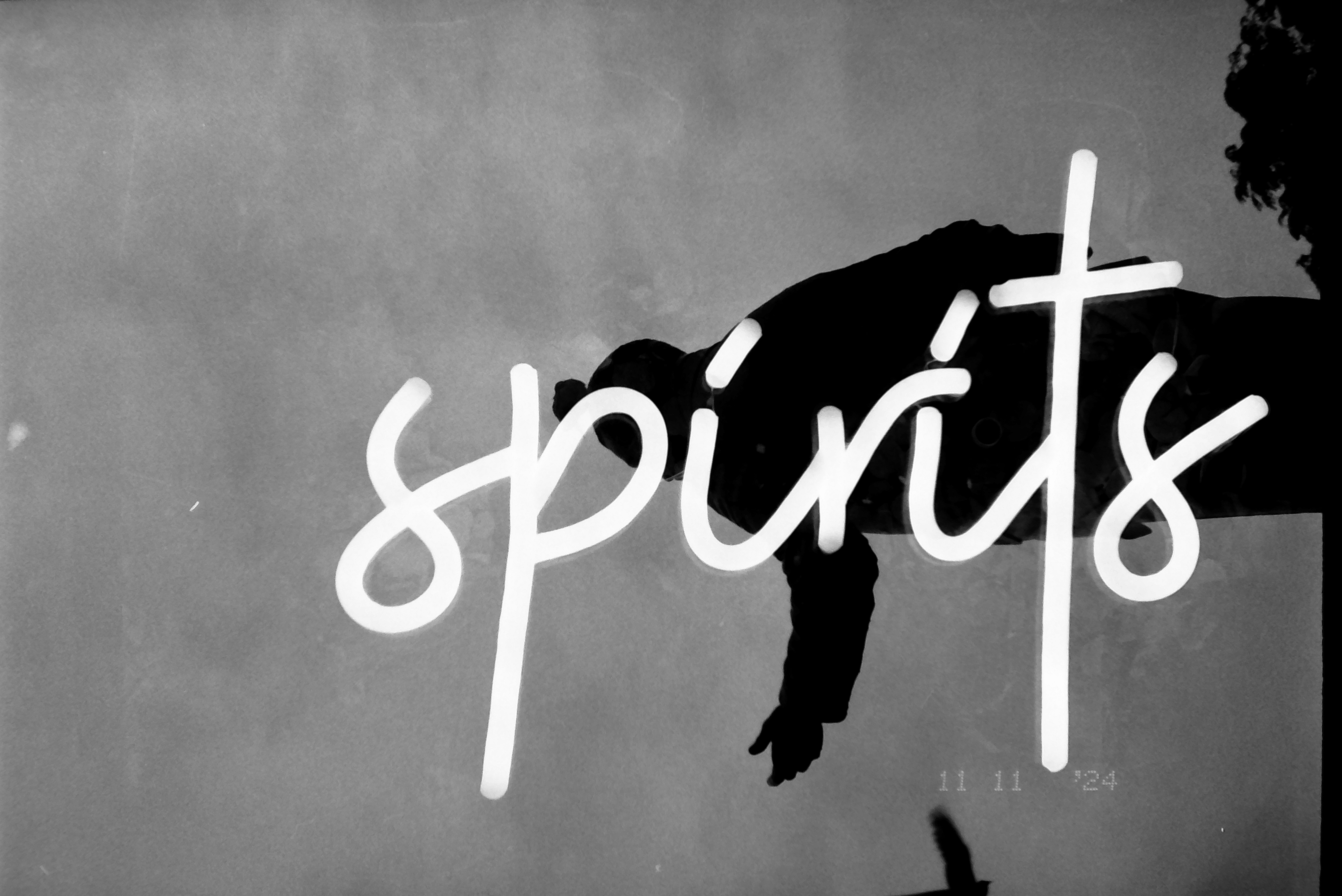 Glowing cursive script spelling 'spirits' crosses a dark, indistinct silhouette against a plain gray background, oriented sideways in the frame.
