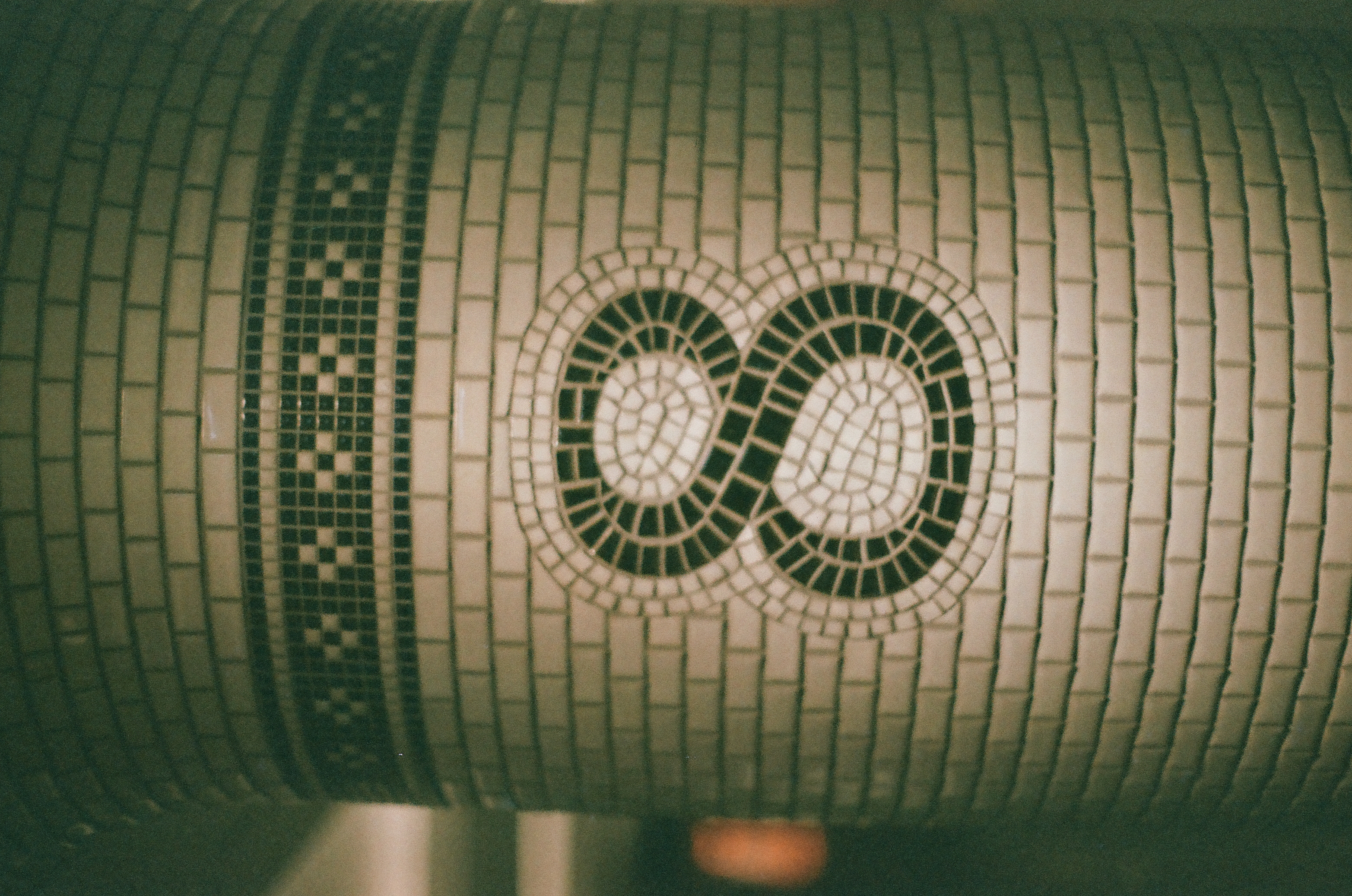 A tiled column displays the numeral eight formed from two connected loops of dark mosaic tesserae beneath a checkered band, with a soft orange light glowing in the lower corner.