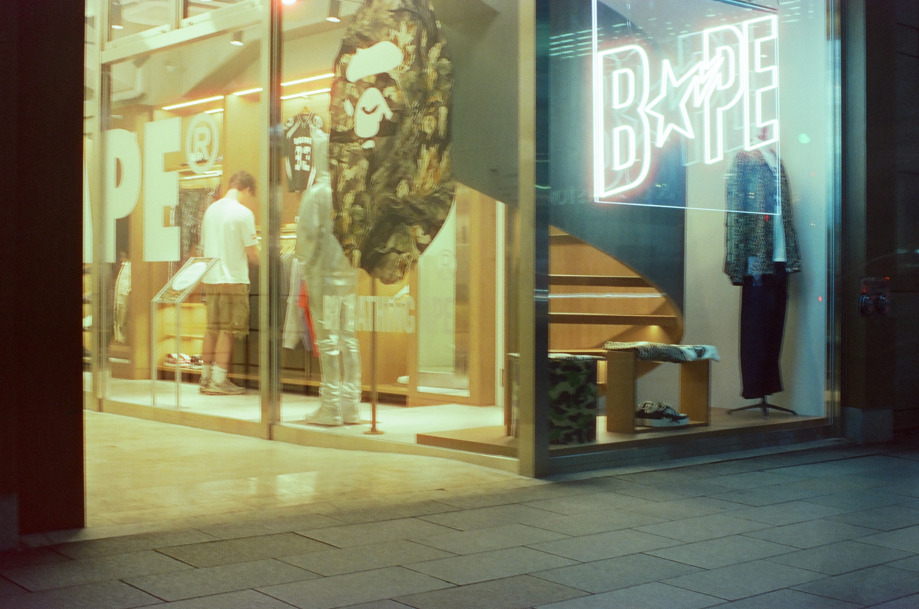 A glass-fronted clothing store lit from inside, with a neon outline sign, a large camouflage-patterned ape-head graphic, two mannequins, and one person standing among the racks.