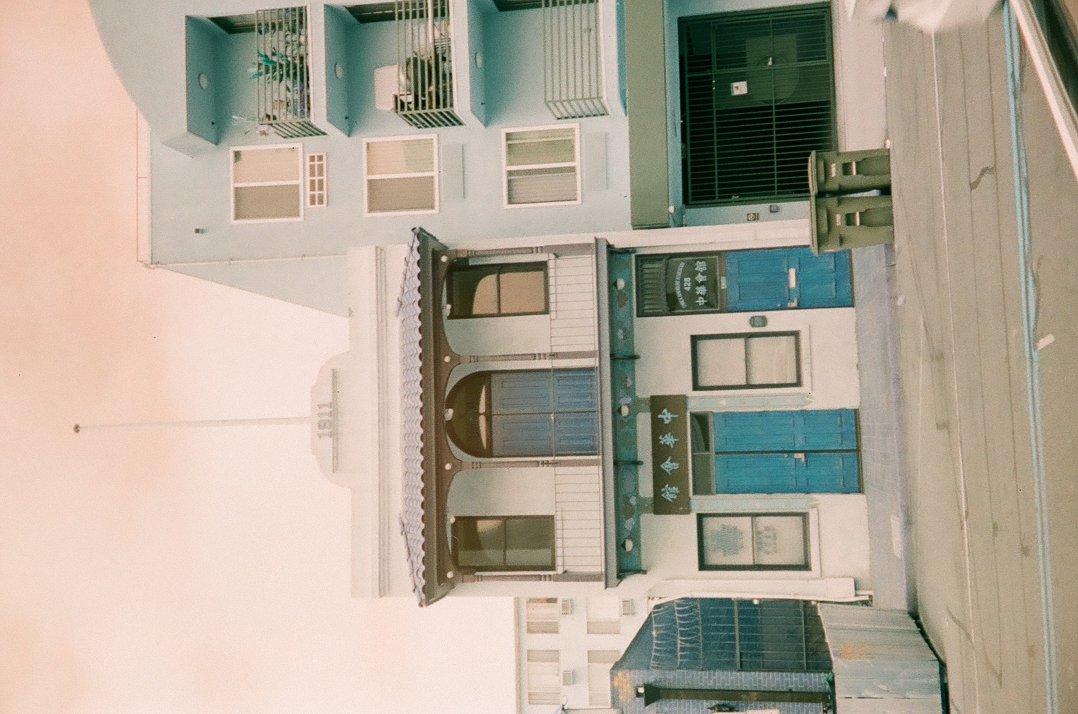 A narrow blue storefront stands between pale neighboring buildings along a quiet sunlit street.
