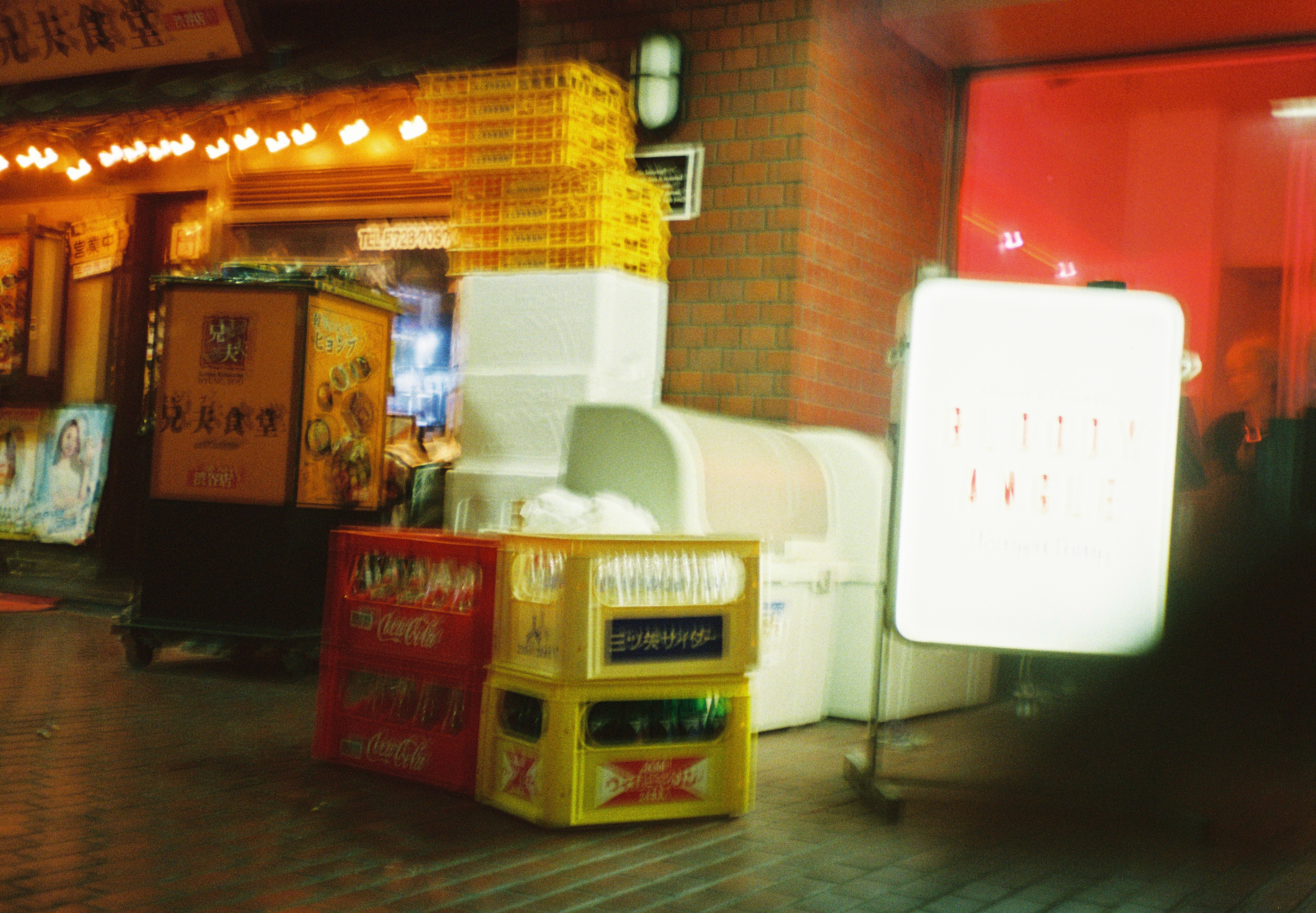 Red and yellow plastic crates and white foam boxes are stacked outside a storefront at night, beside a glowing white sign.
