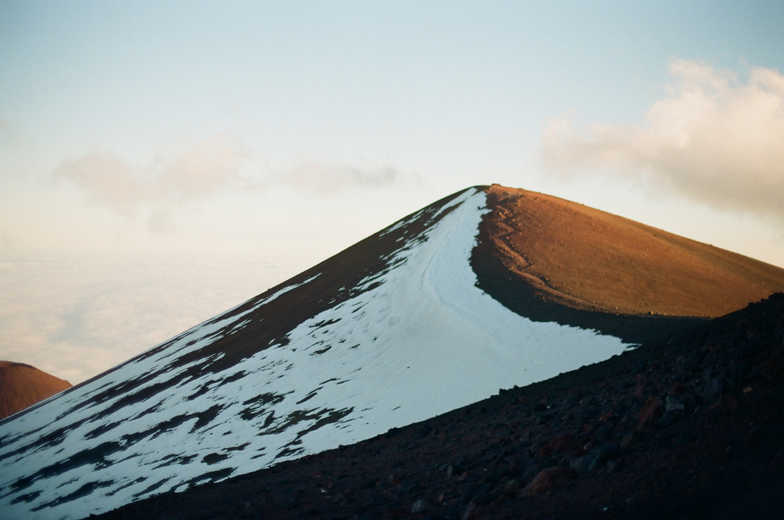 A long white snowfield follows the steep edge of a rust-colored ridge beneath soft clouds.