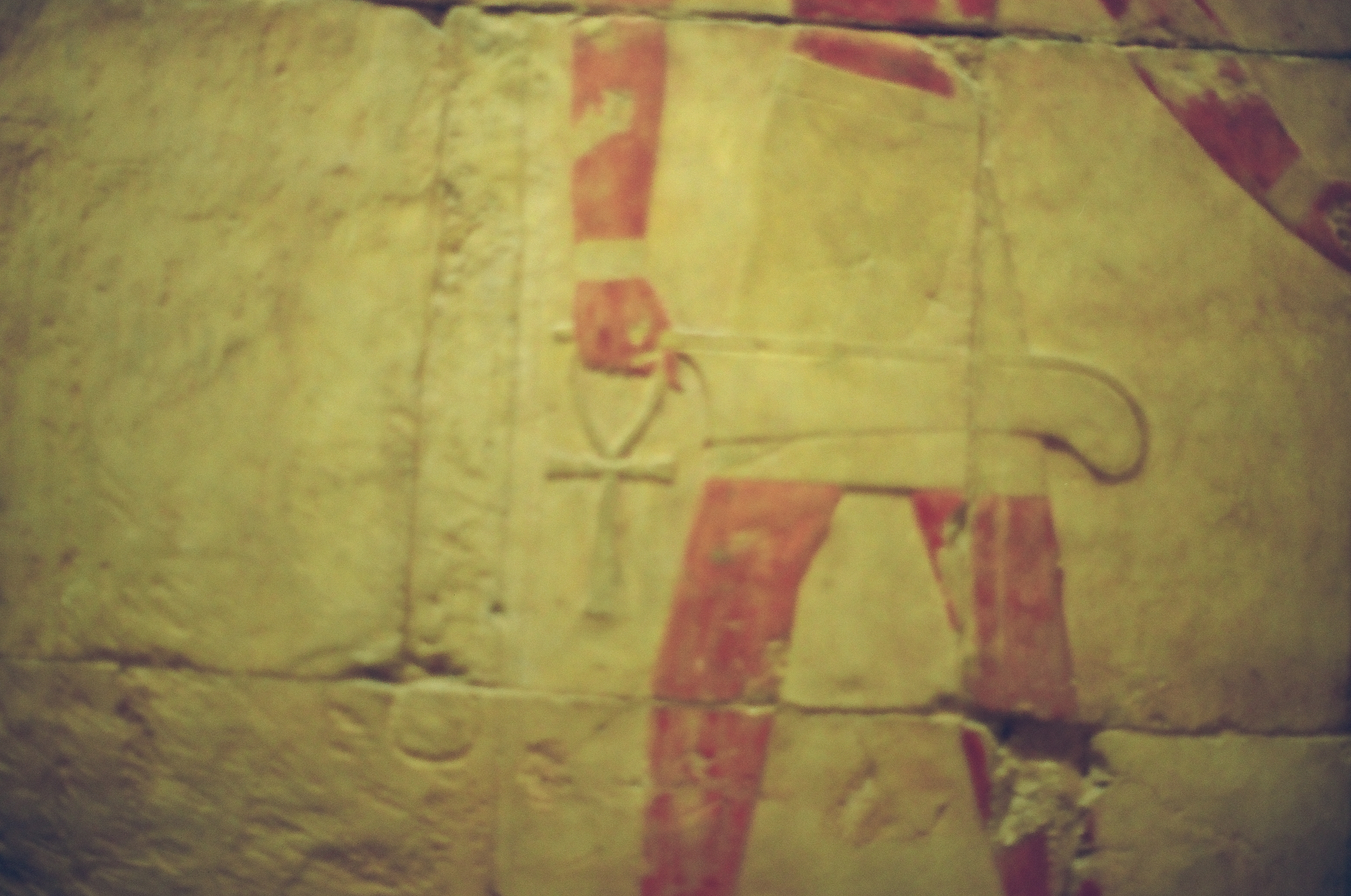 A carved stone relief shows the red-painted striding legs of a figure whose hand holds an ankh-shaped loop, with block joints crossing the surface.