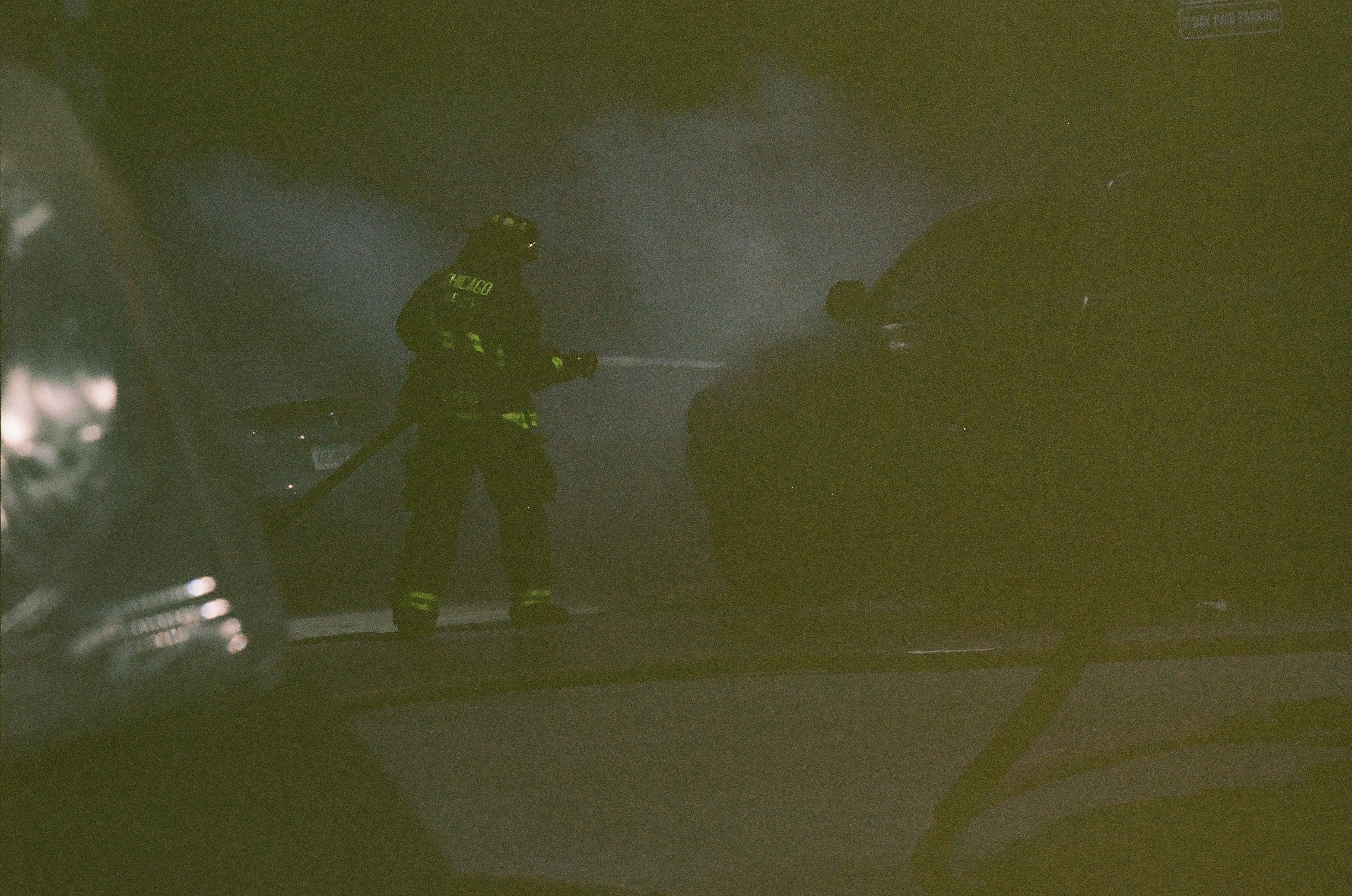 A firefighter directs a hose through heavy smoke beside dark vehicles and another shadowed figure.