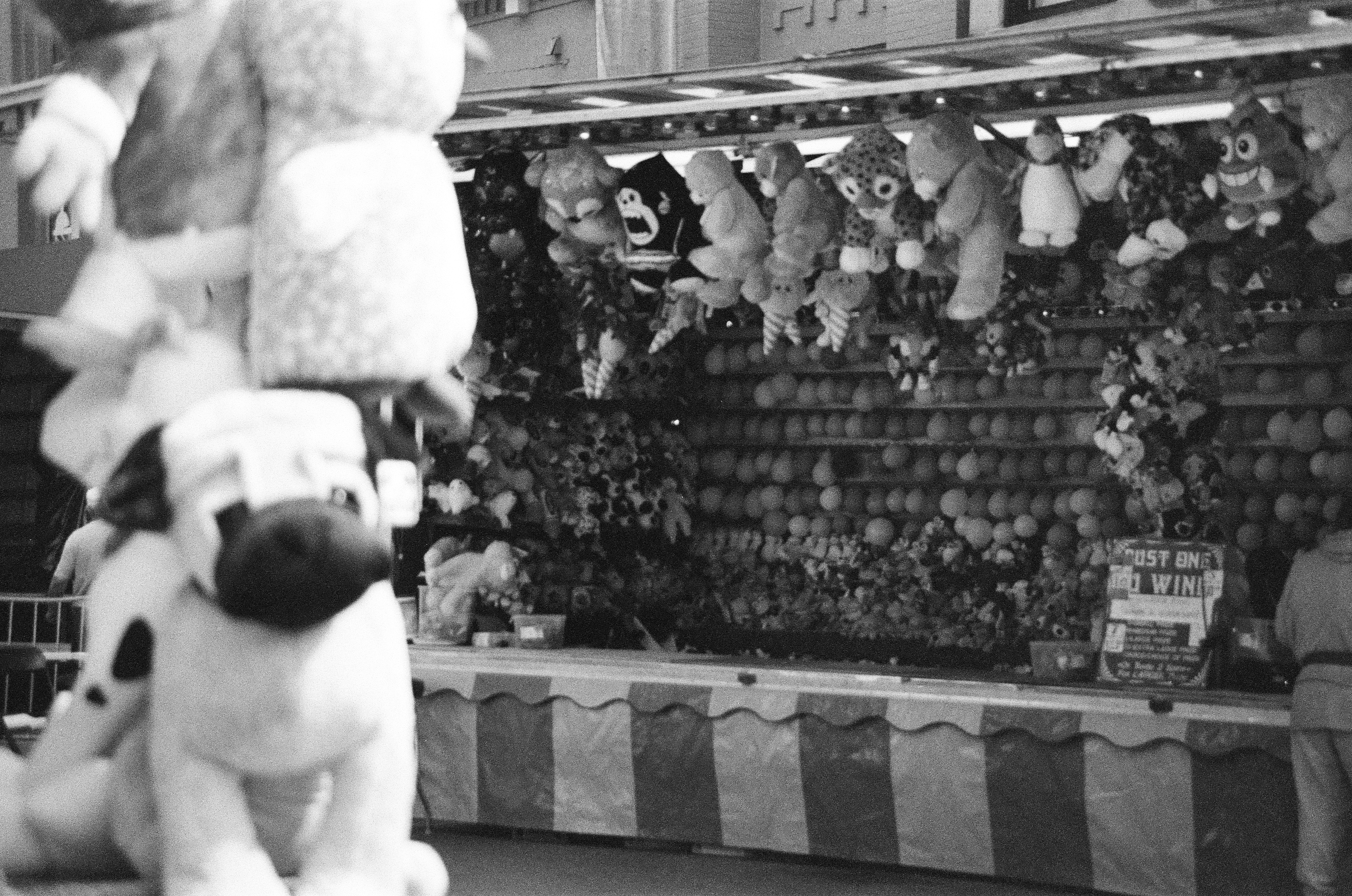 Blurred plush animals hang in the foreground before a crowded booth packed with prizes and circular targets.