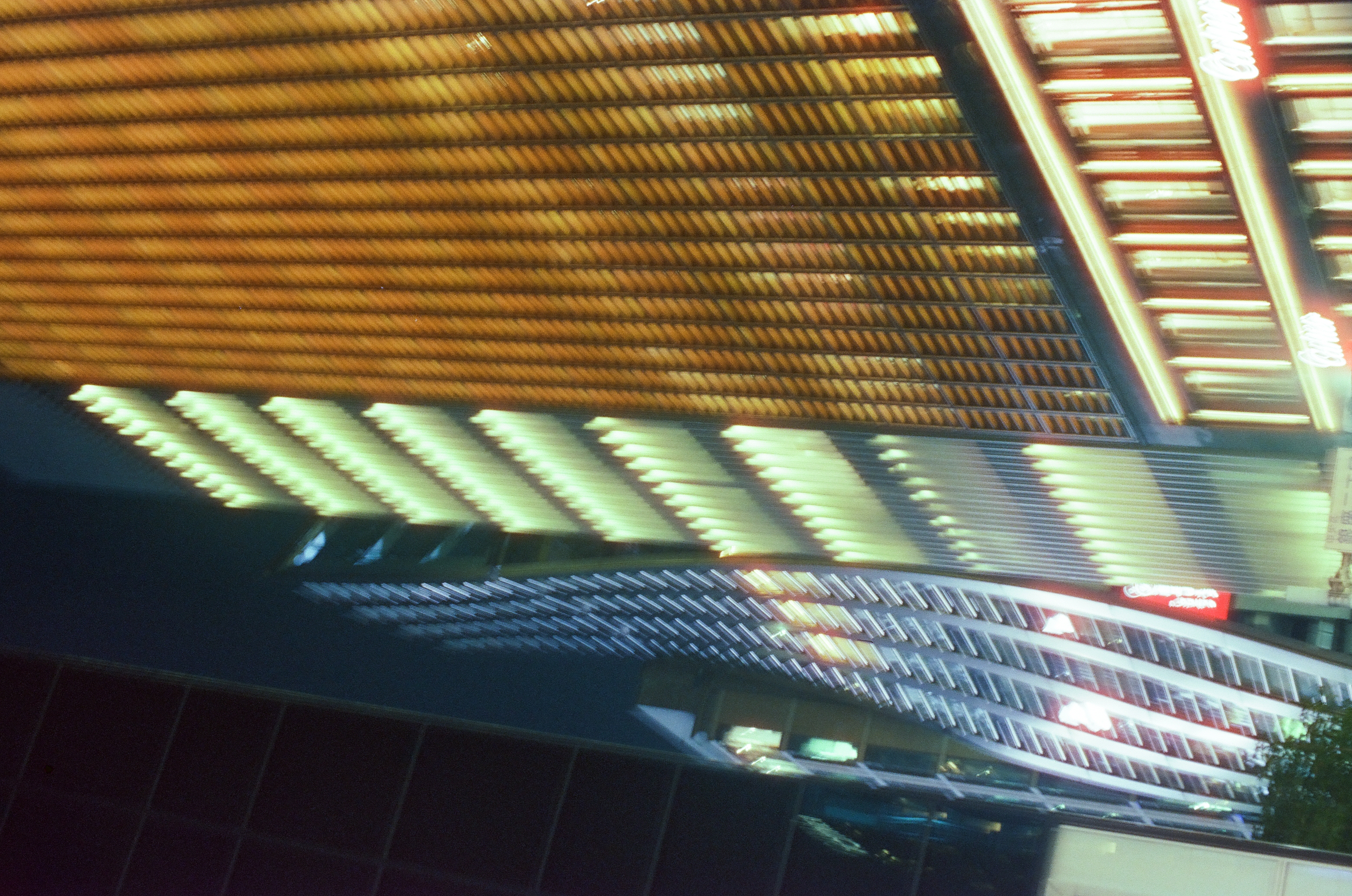 A tilted view of building facades with rows of lit louver-like slats, amber below and pale green and blue above, beneath a dark gridded facade at upper right.