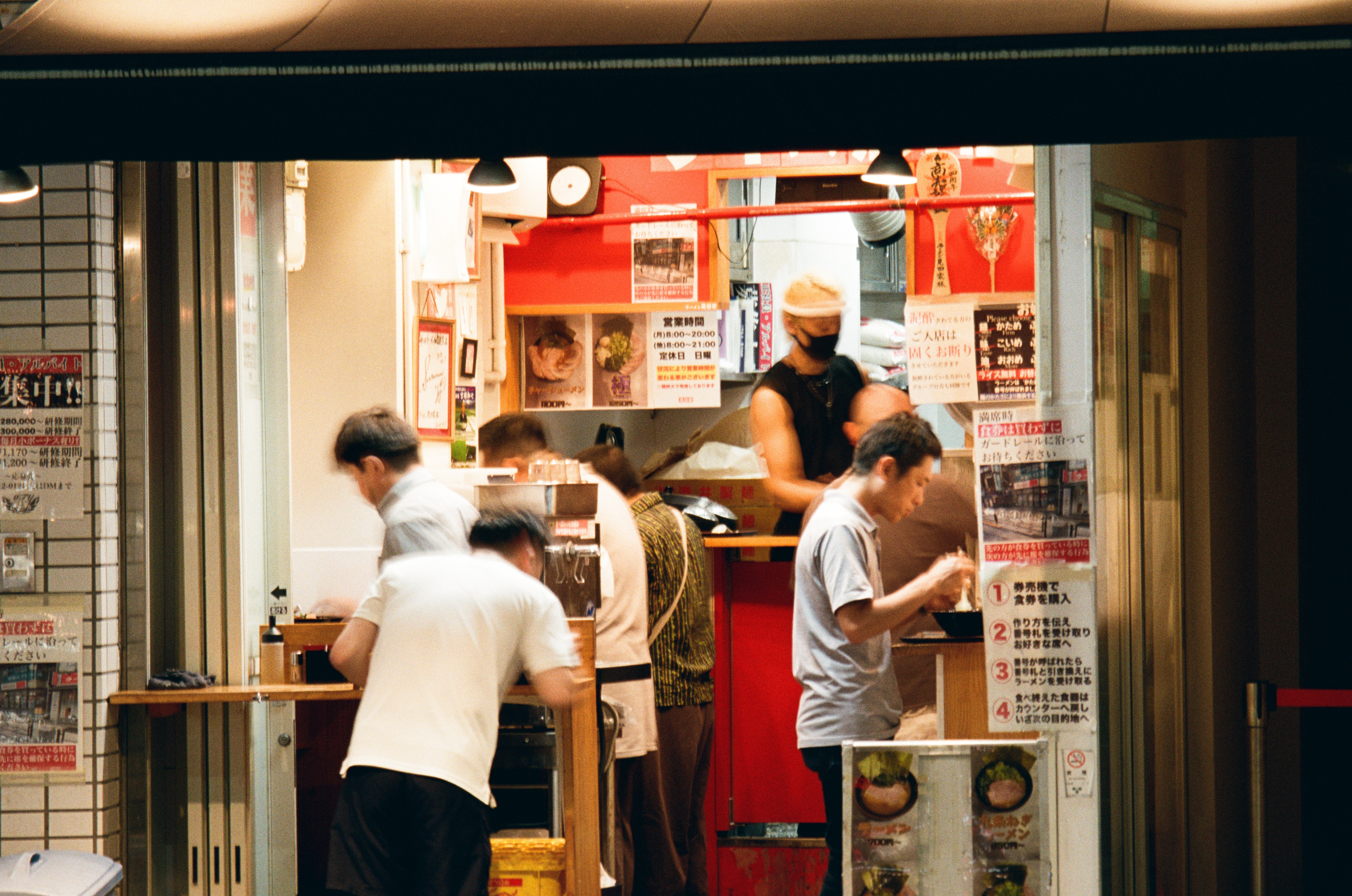 Cooks and customers in white shirts crowd an open-fronted ramen counter at night, its red walls papered with handwritten menus and notices.