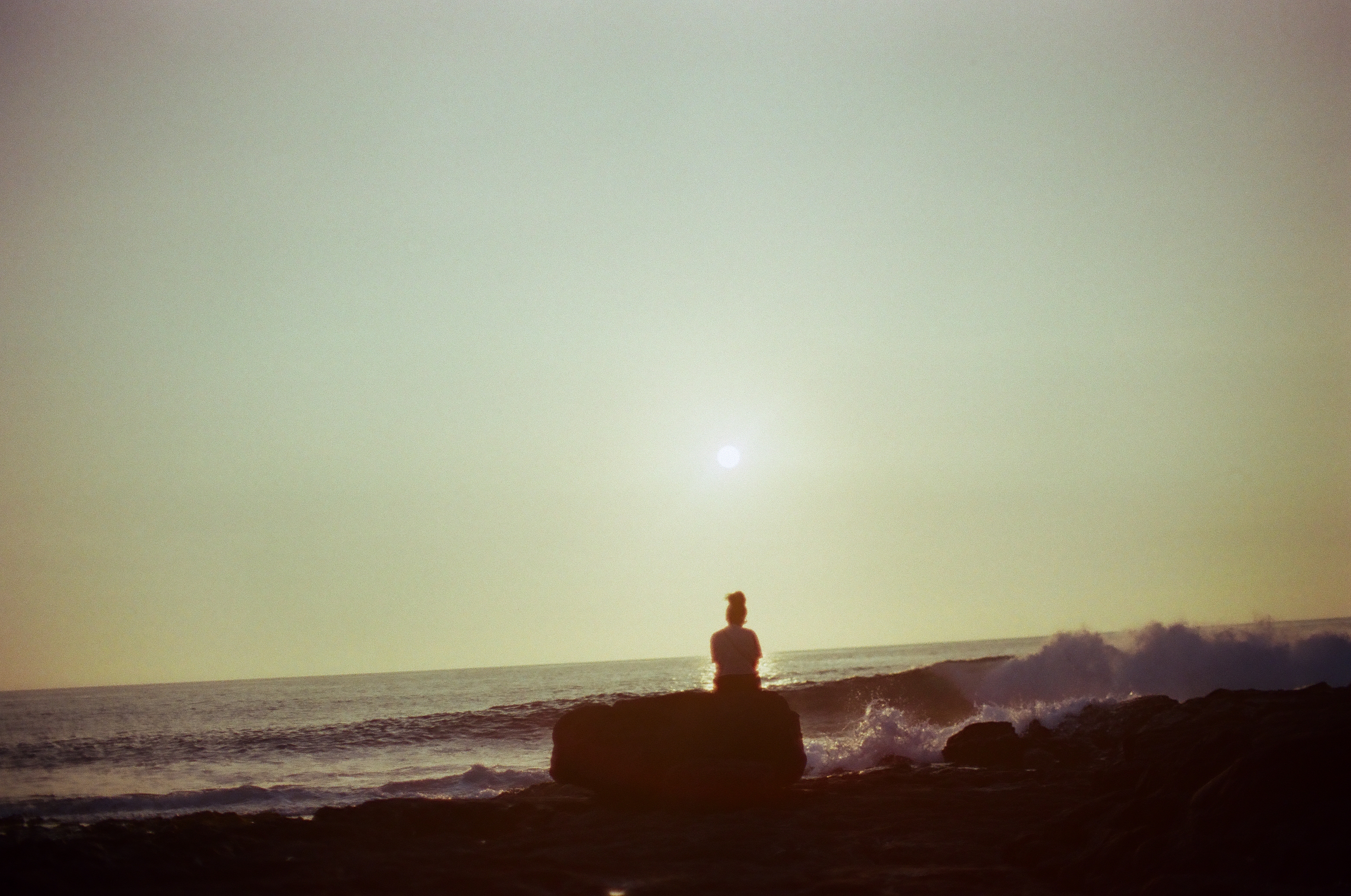 A small seated figure faces the ocean from a dark rock while a low sun glows through sea haze.