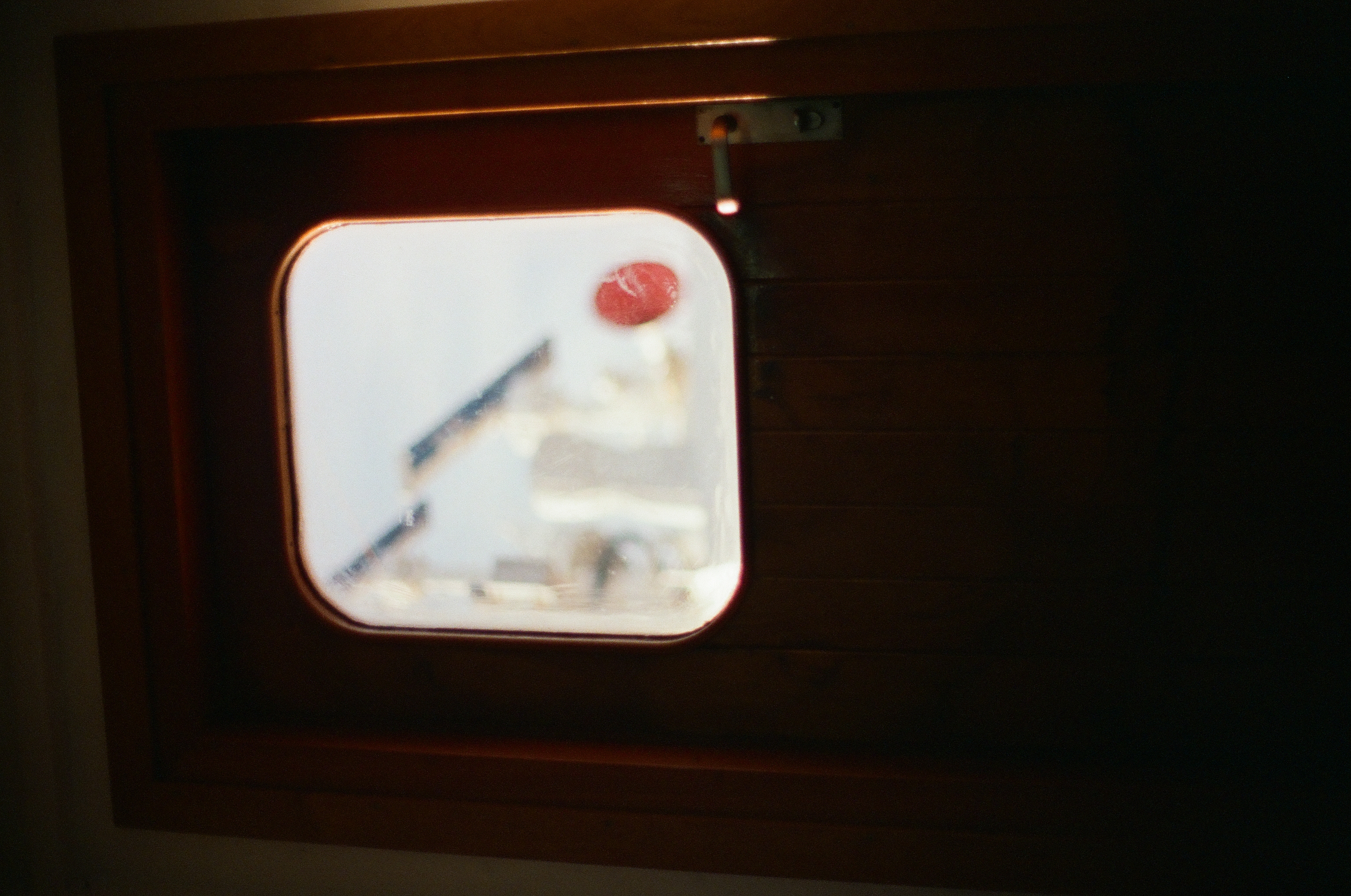 A rounded window set in a varnished wood door frames a blurred red shape and pale deck structure outside, beside a small metal latch.