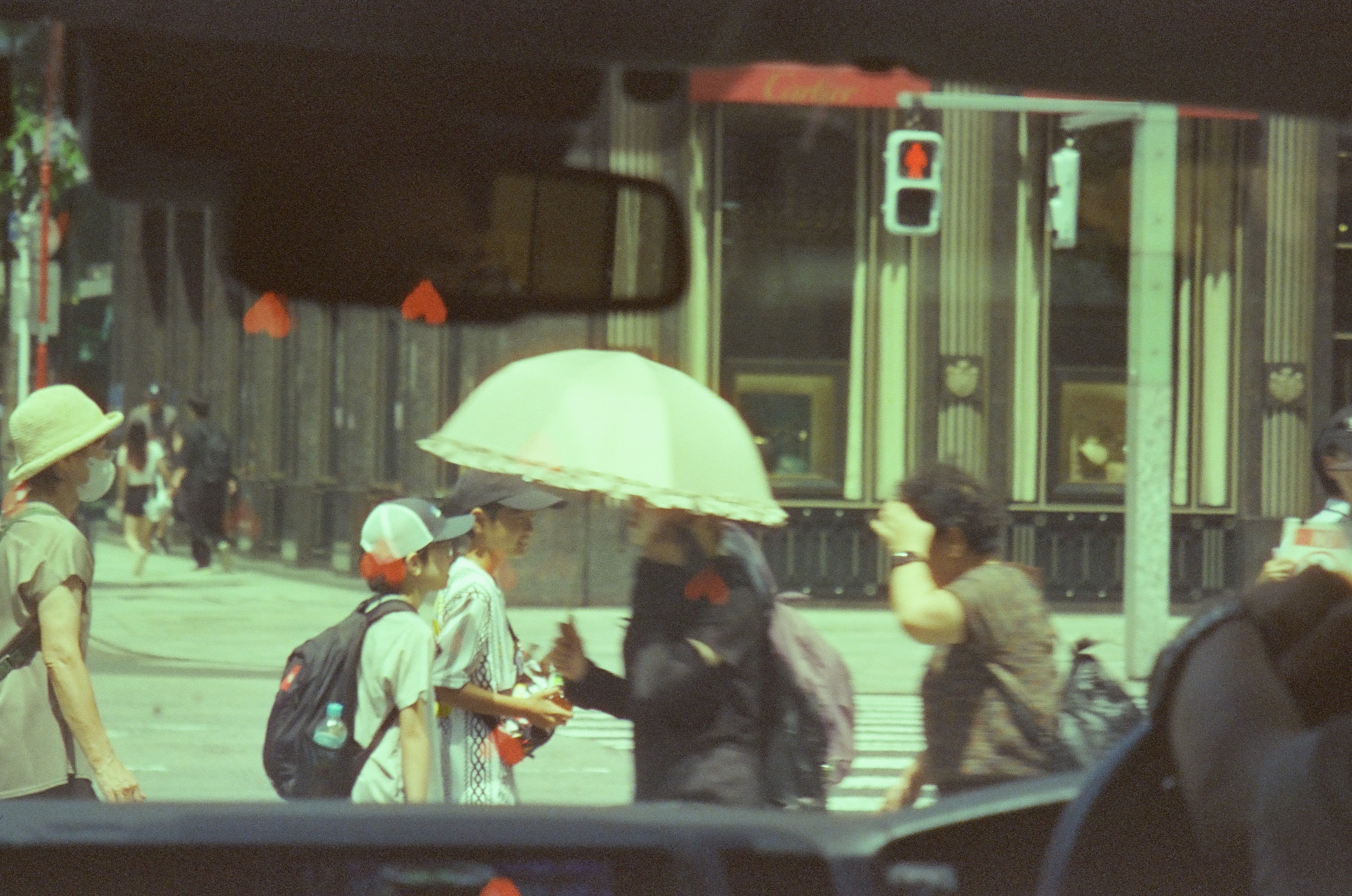 Pedestrians with pale umbrellas cross a bright street beneath small red and white heart marks.
