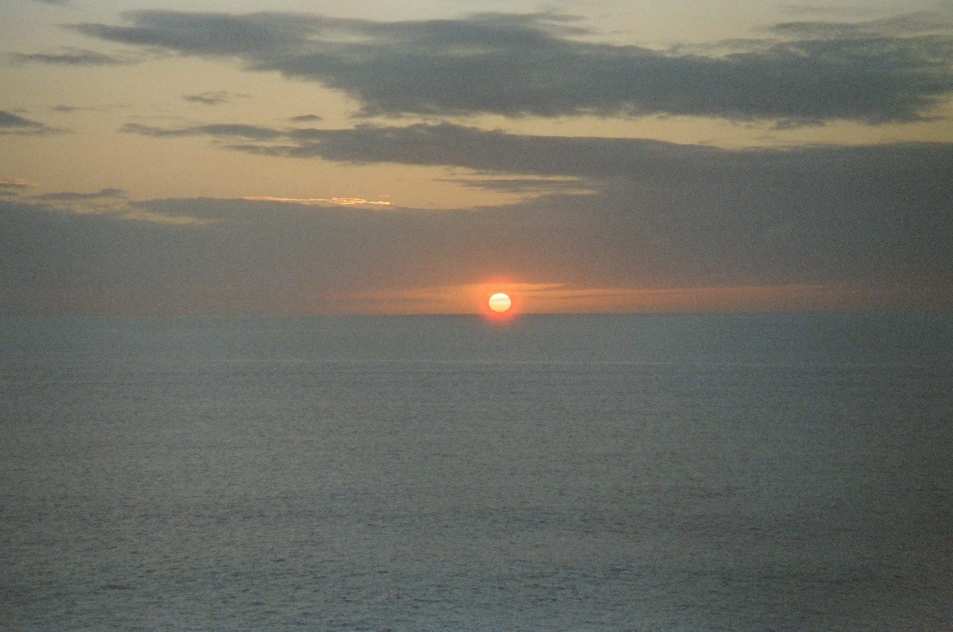 A small red-orange sun sits on the sea horizon beneath a dark band of clouds, with calm gray-blue water below.