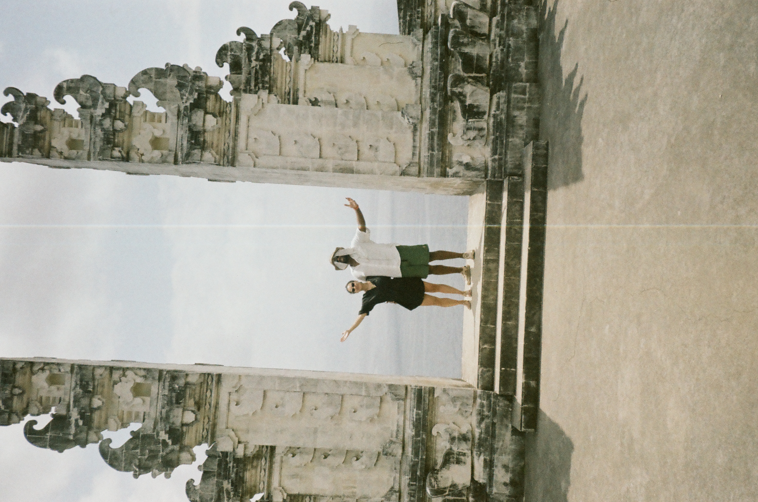 Two people each extend an outer arm while standing between carved towers, crossed by a pale vertical line near the center.