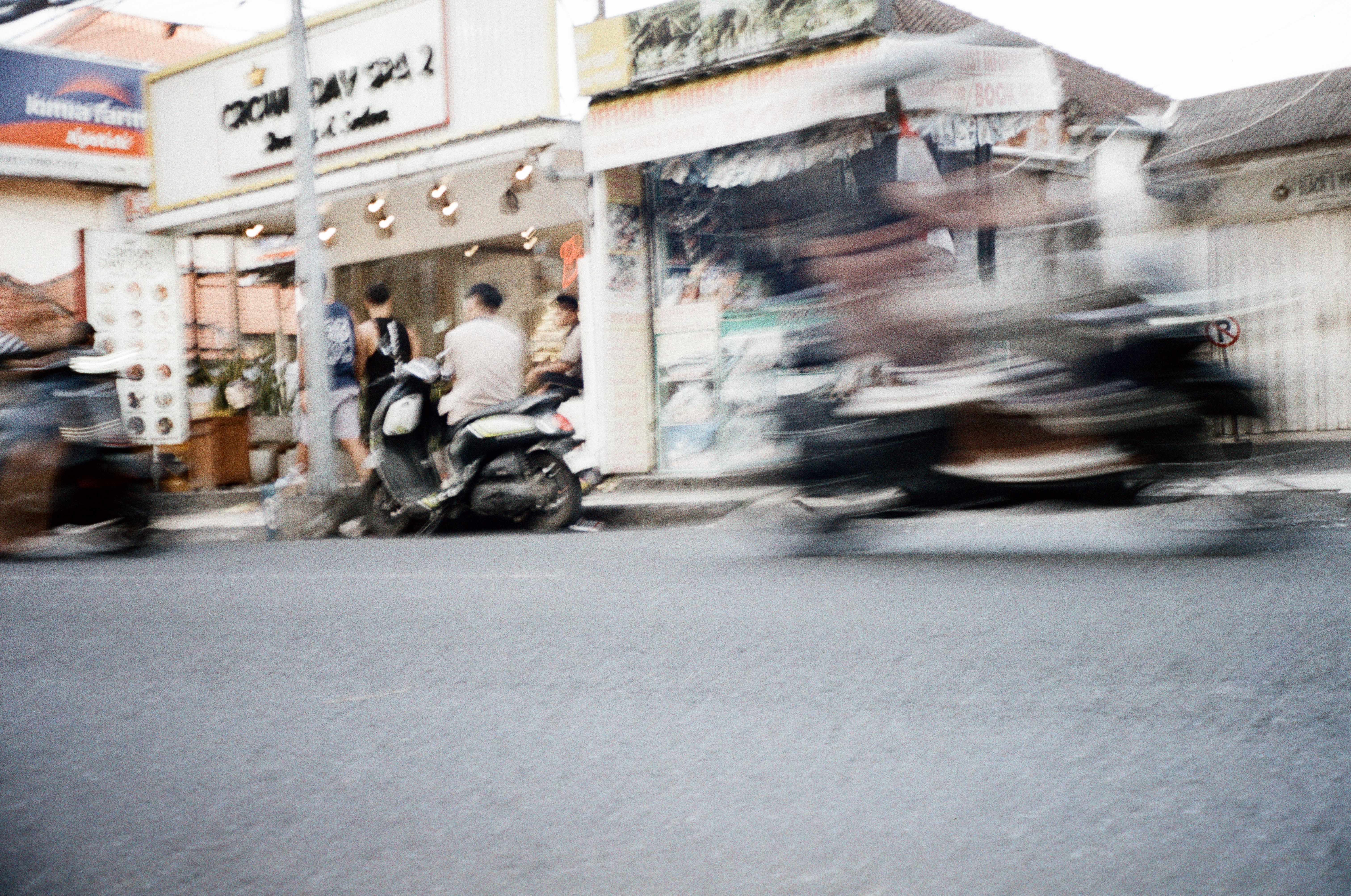 Two scooters blur across the foreground of a street lined with shops, where people stand near a parked scooter under lit signs.