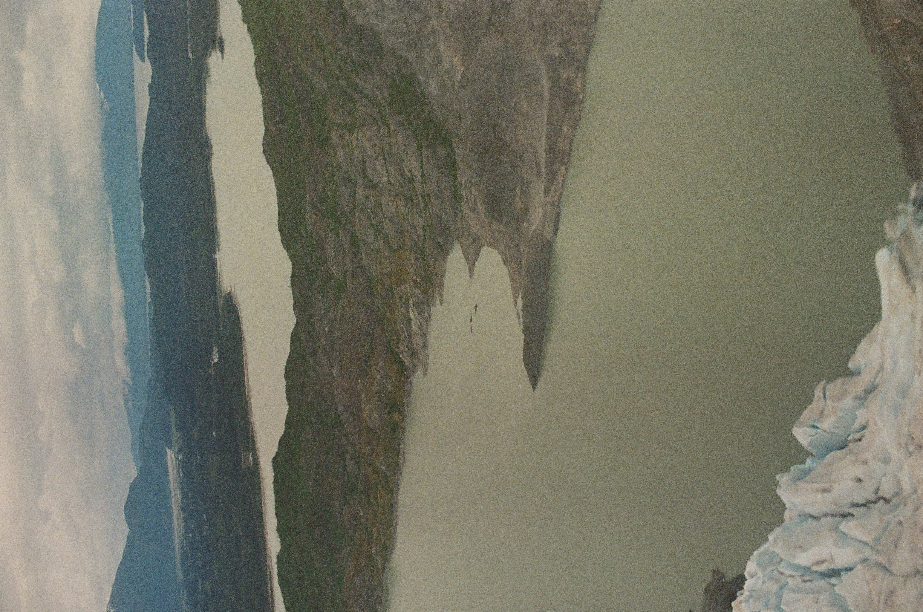 Two muted green lakes lie between dark ridges beyond a rough blue-white edge of glacier ice.