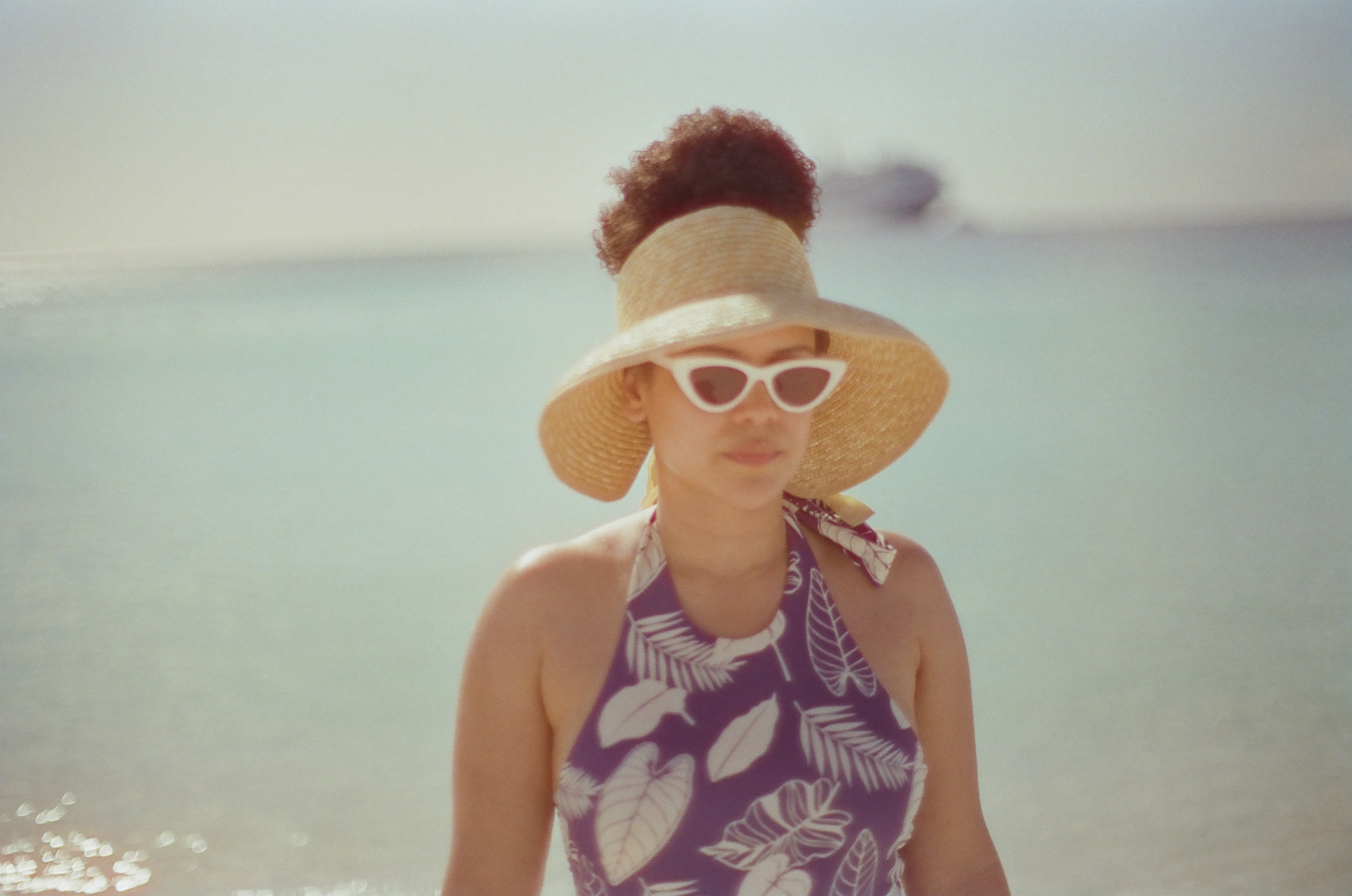 A person wearing a wide straw visor and white sunglasses stands before bright water in direct afternoon light.