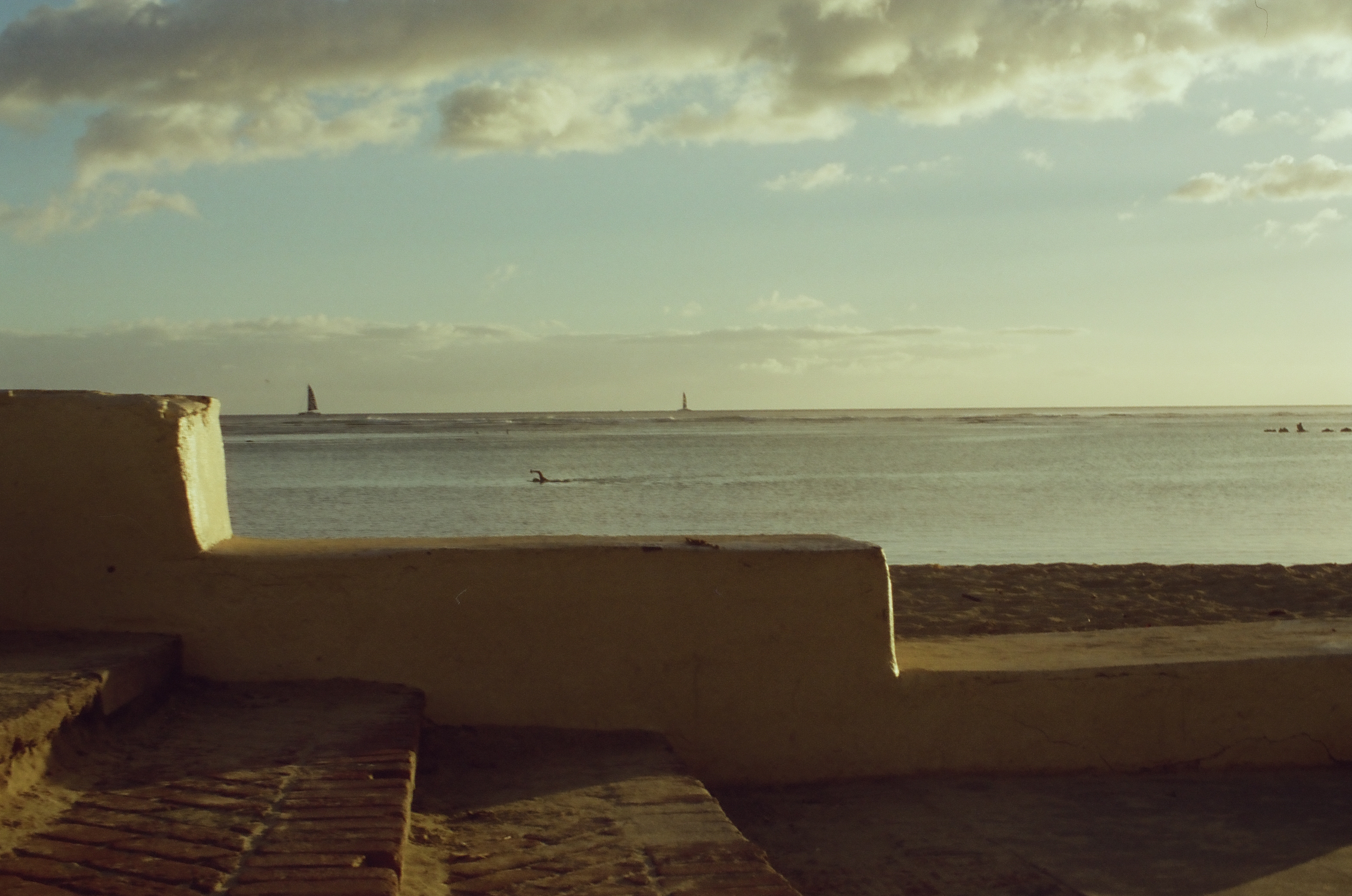 A low pale sea wall borders calm water beneath warm clouds with tiny sailboats scattered along the horizon.