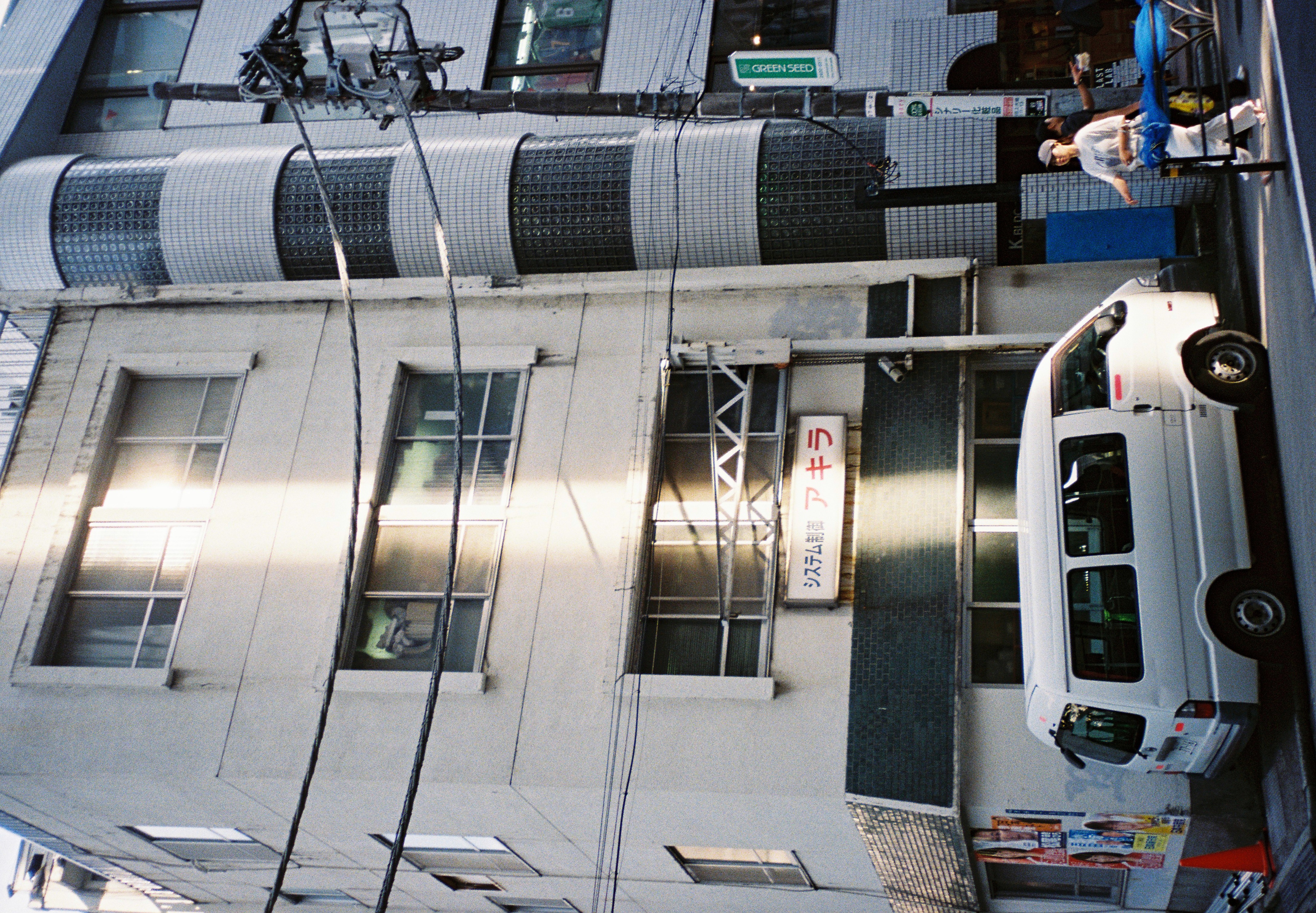 The full height of a tiled office building, joined by a rounded glass-block tower, rises beside a parked van as a pedestrian crosses the street.