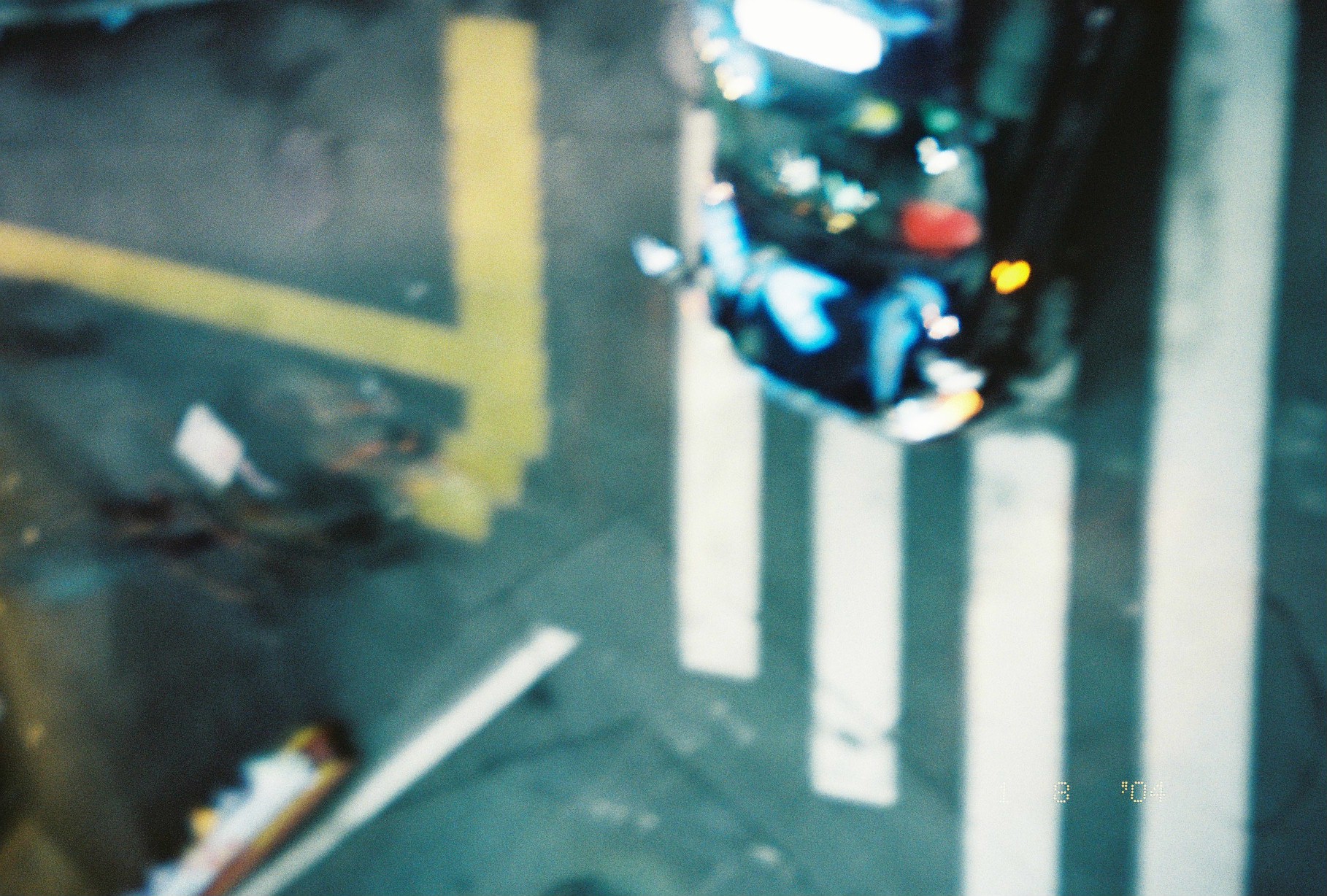 Seen from an overpass, a dark car with bright headlights crosses broad white crosswalk stripes beside a curved yellow lane marking.