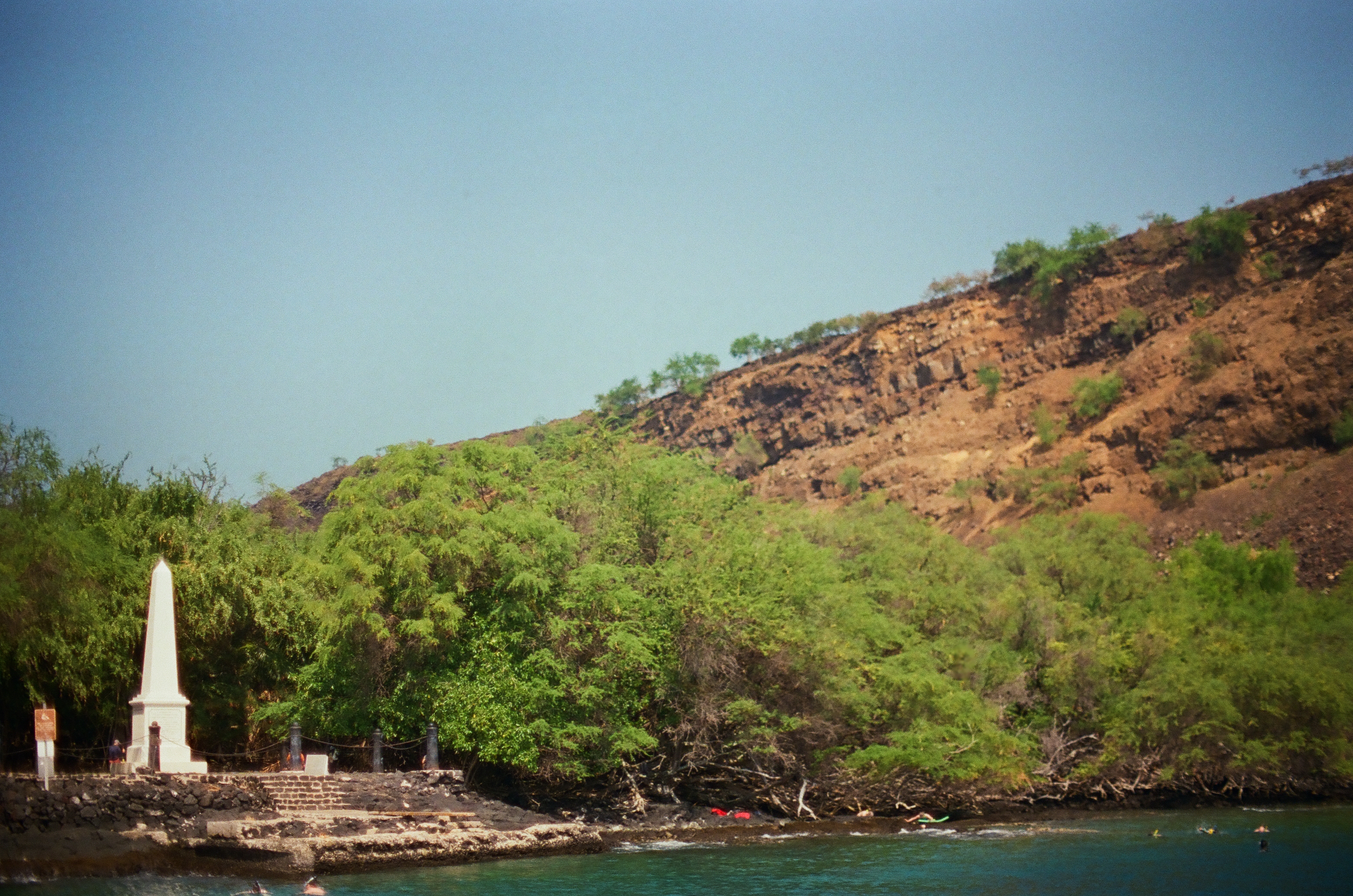 A slender white monument rises at the edge of a green cove where dark rocks meet calm blue water.