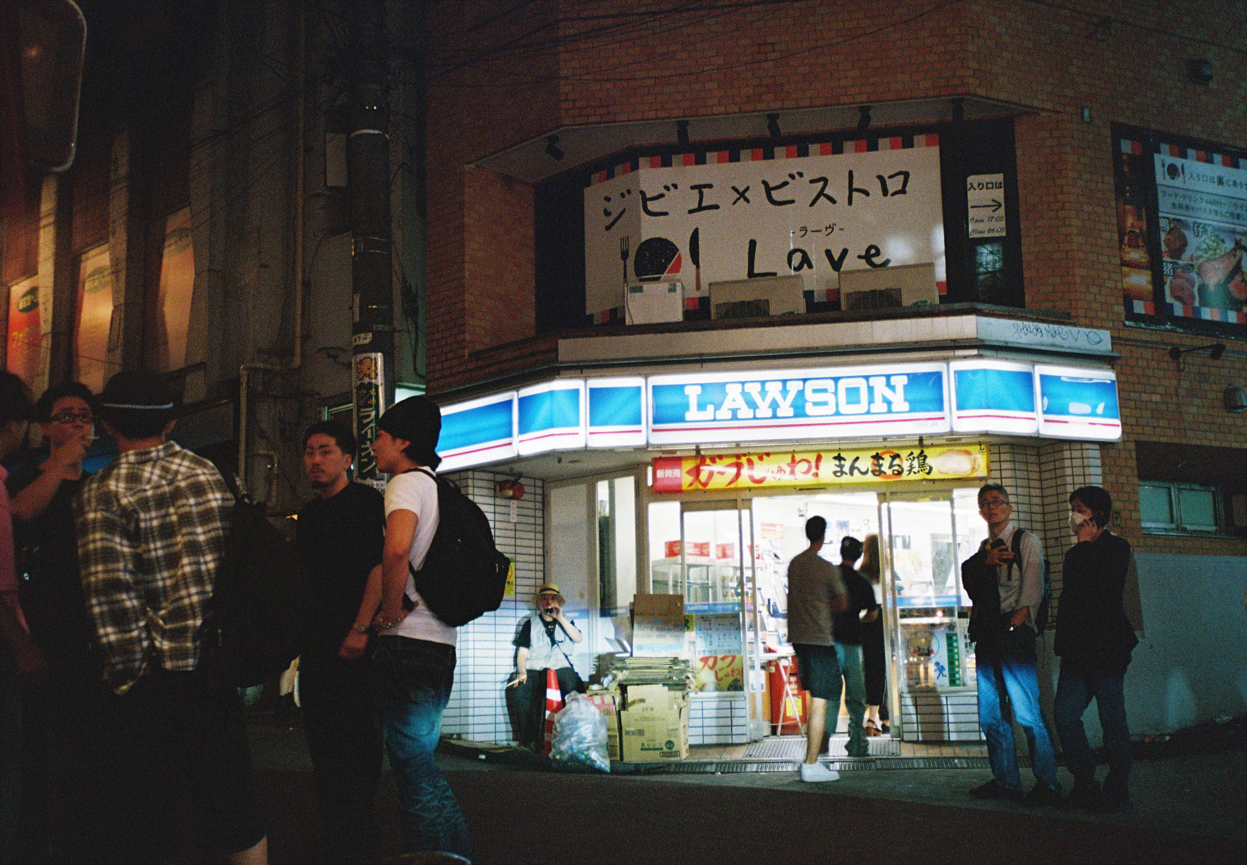 Several people, some wearing backpacks, stand near a lit storefront entrance where boxes and a traffic cone are piled beside the door.