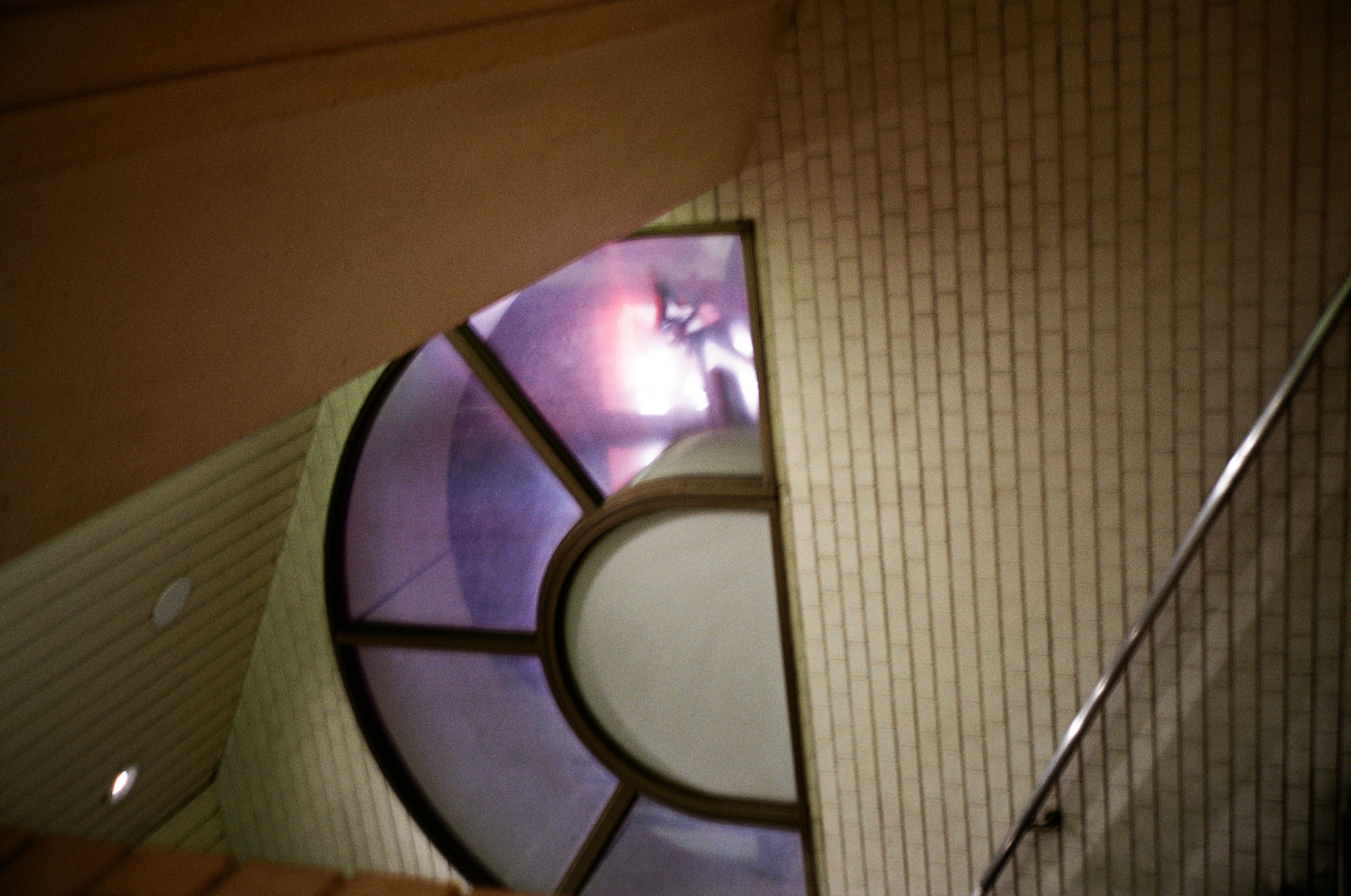 A large fan-shaped arched window glows violet against cream tiled walls in a stairwell, a metal handrail crossing the lower frame.