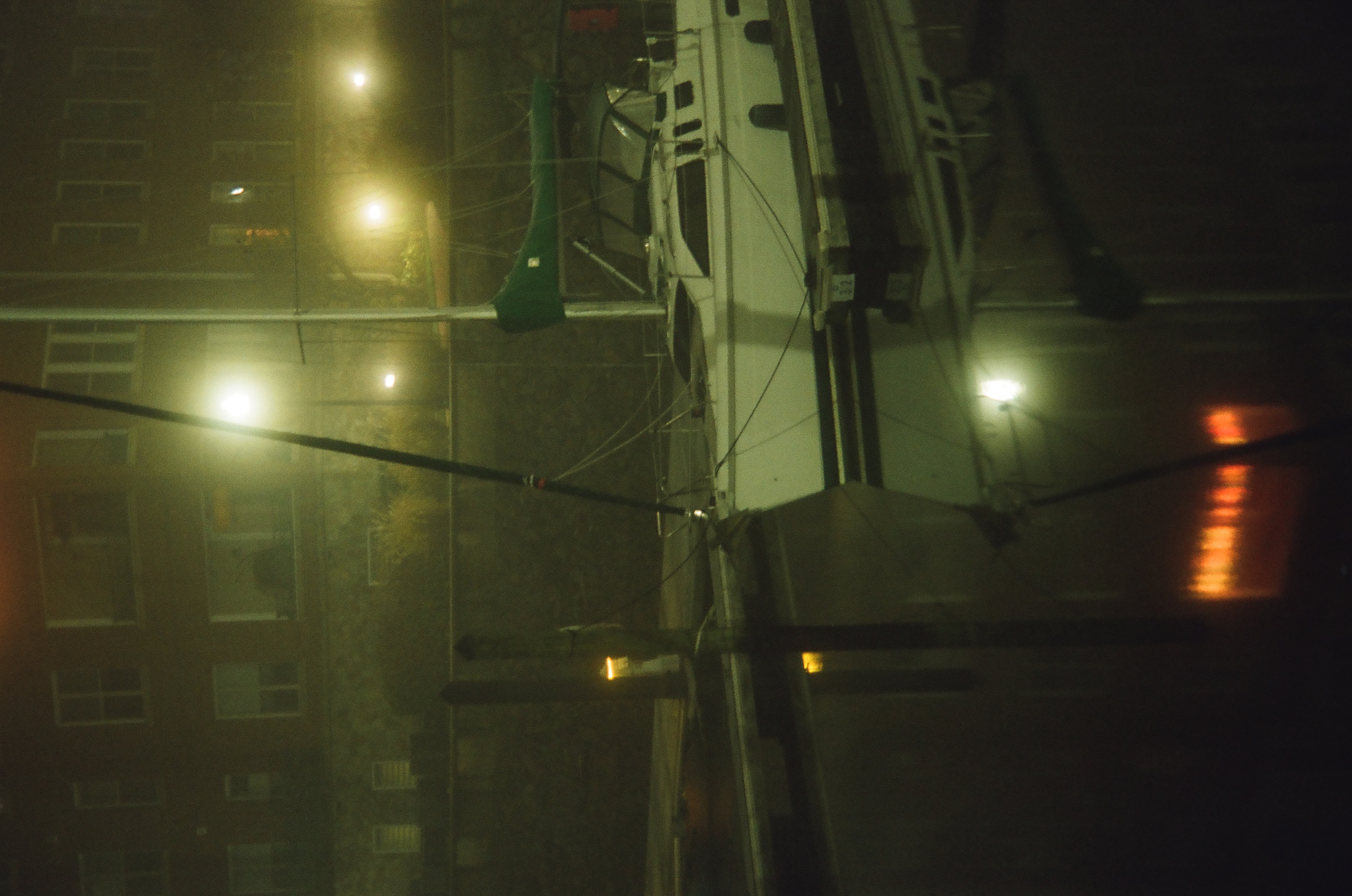 A white sailboat with a furled green sail cover sits moored beside a bulkhead in front of a lit building, its mast mirrored in still water.