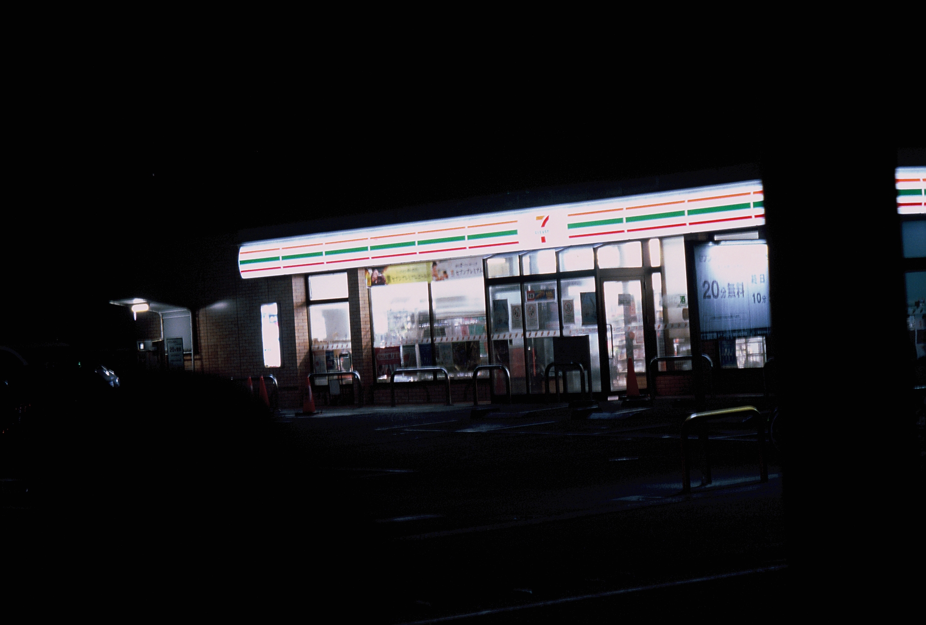 A brightly lit convenience store with red, green, and orange striped signage stands alone against a completely dark street at night.