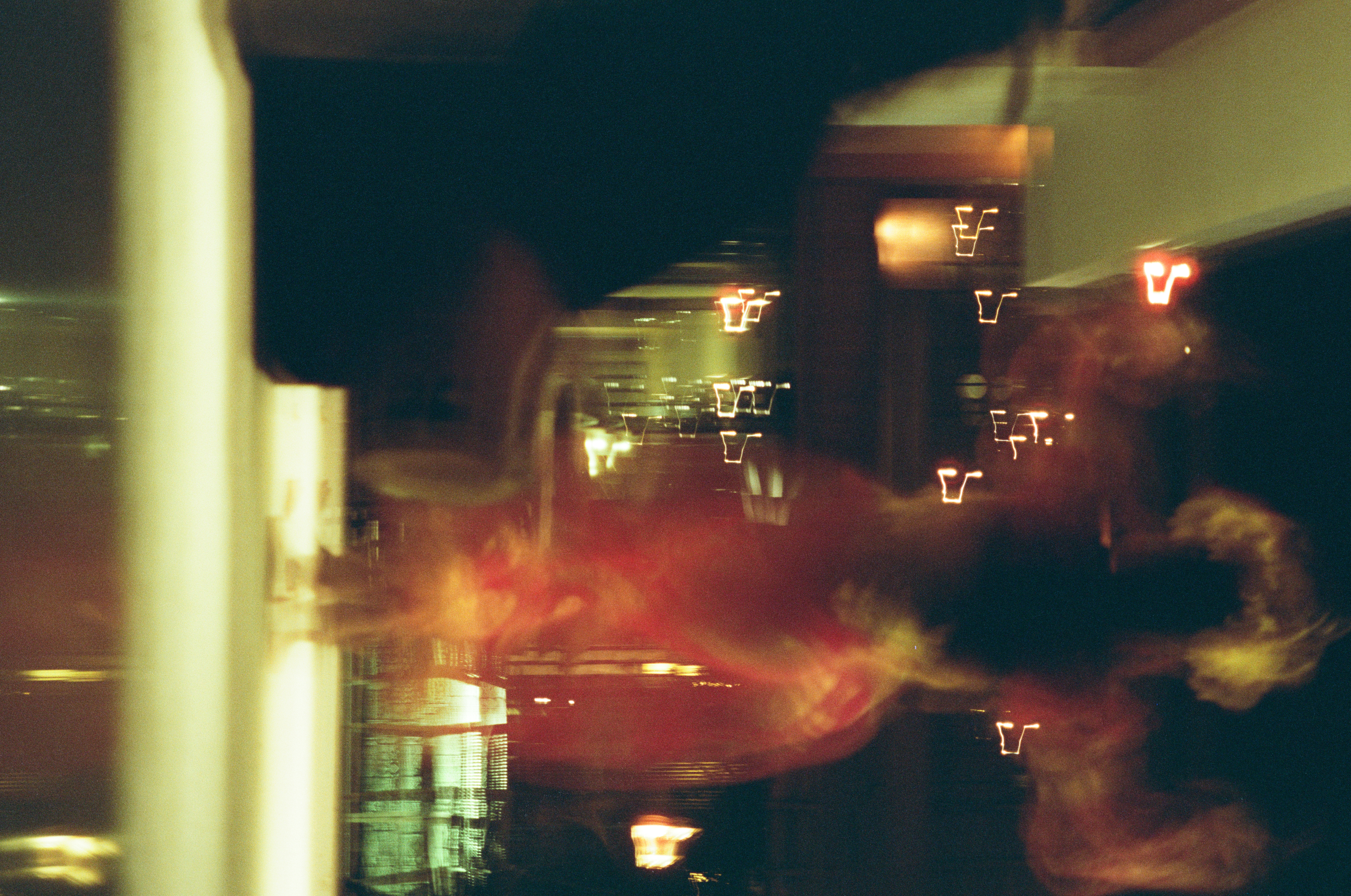 A blurred red-and-gold figure with a large gold head adornment raises both hands amid streaked lights, above a pale floor and dark reflective band.