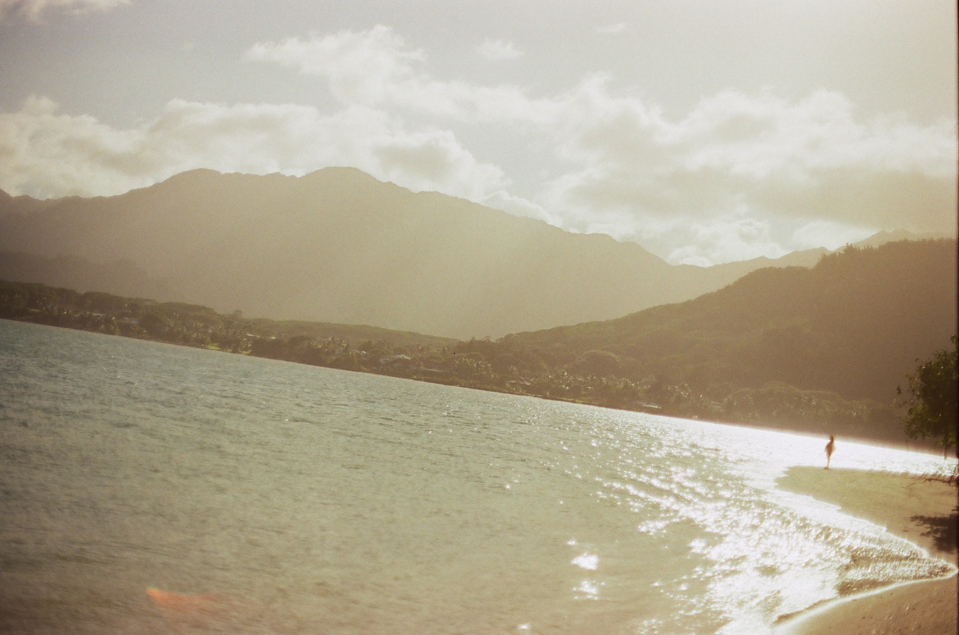 A solitary silhouetted figure walks along a sunlit shoreline beneath hazy backlit mountains and thick clouds, with an orange lens flare in the foreground water.