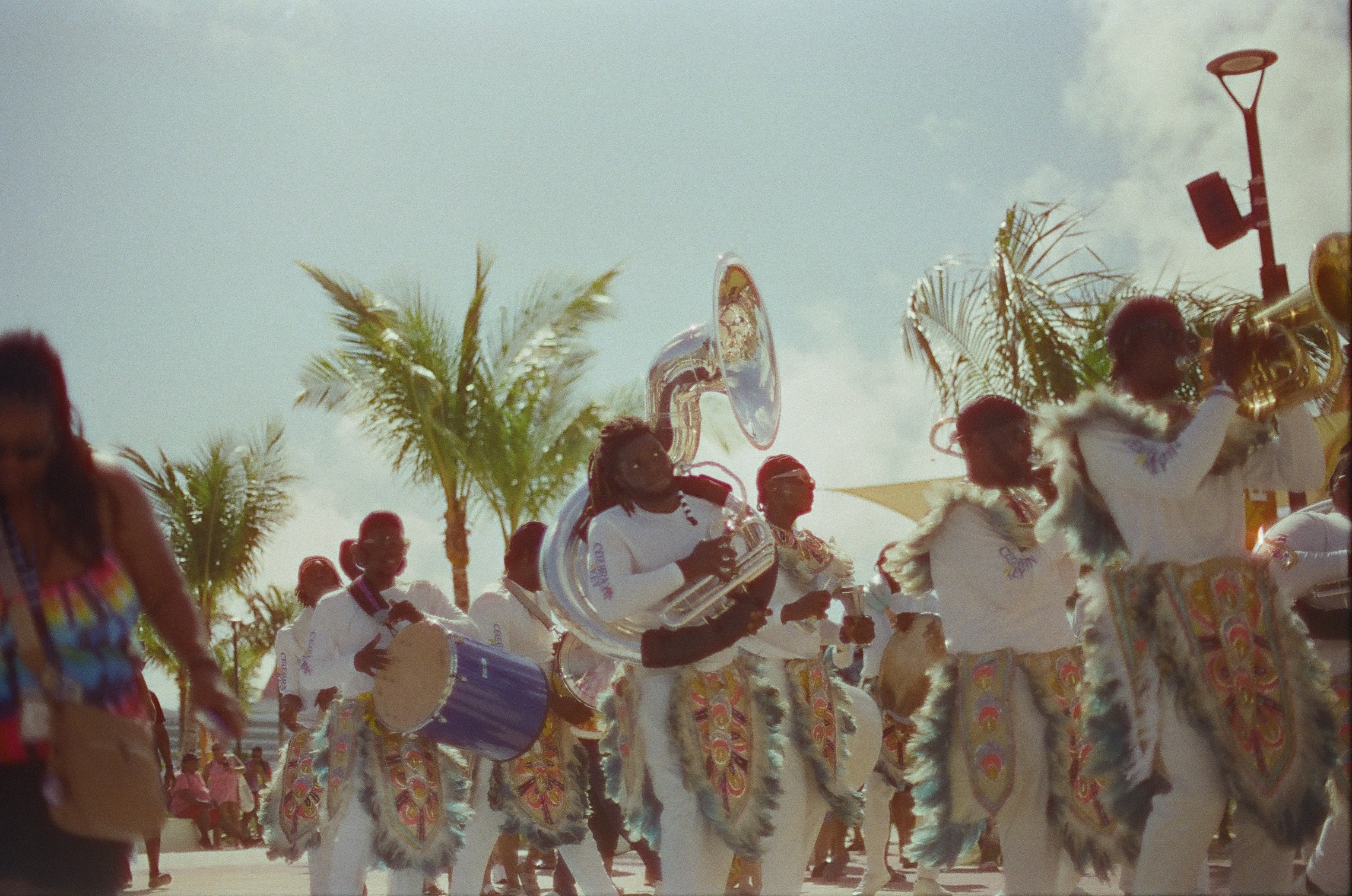 Musicians in white outfits with feathery trim and decorated panels march past palm trees, one carrying a large silver sousaphone and another a blue drum.