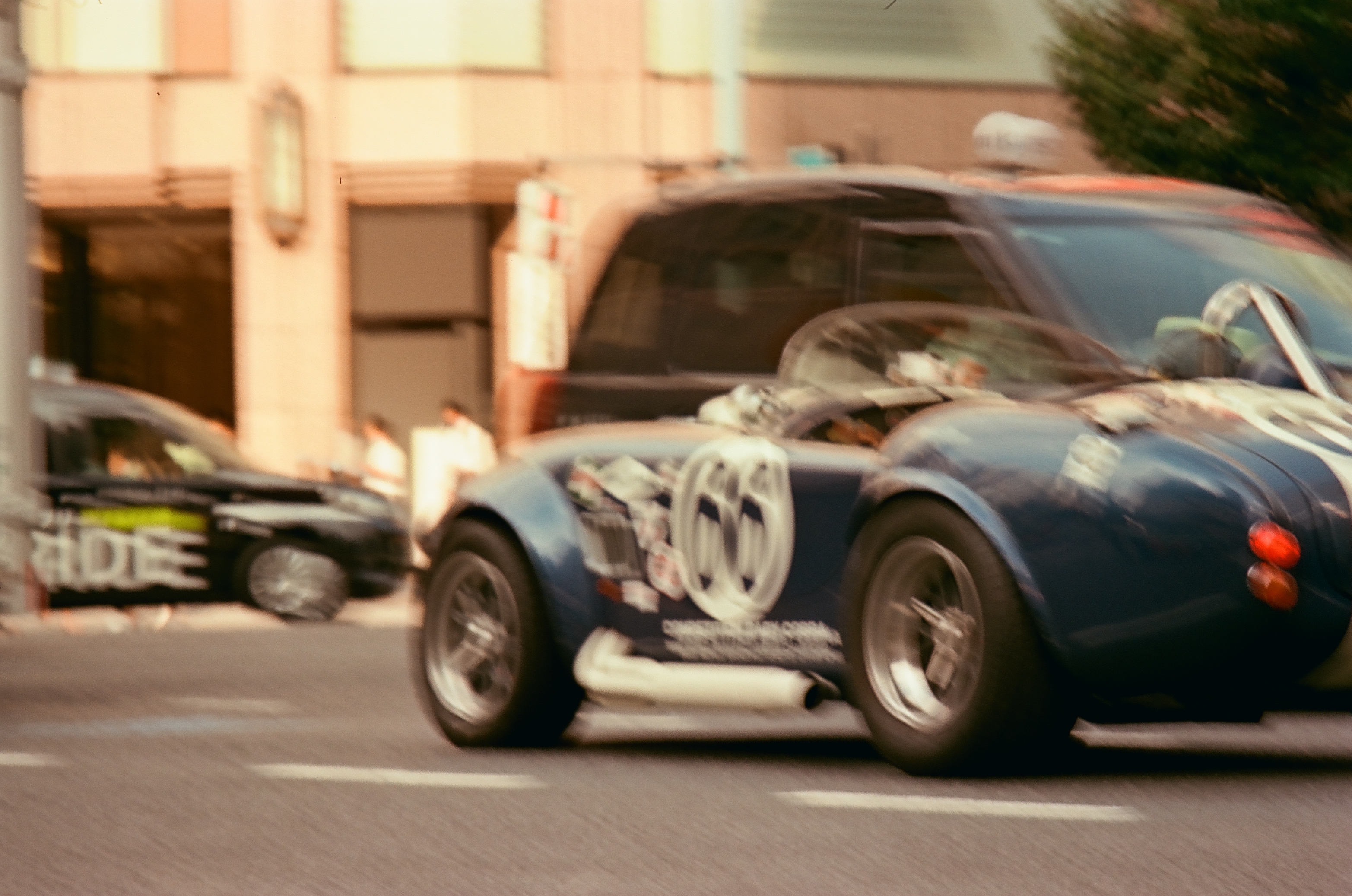 A dark blue open-top sports car with white stripes, a round door number, and side exhaust pipes blurs past a building and a black car.