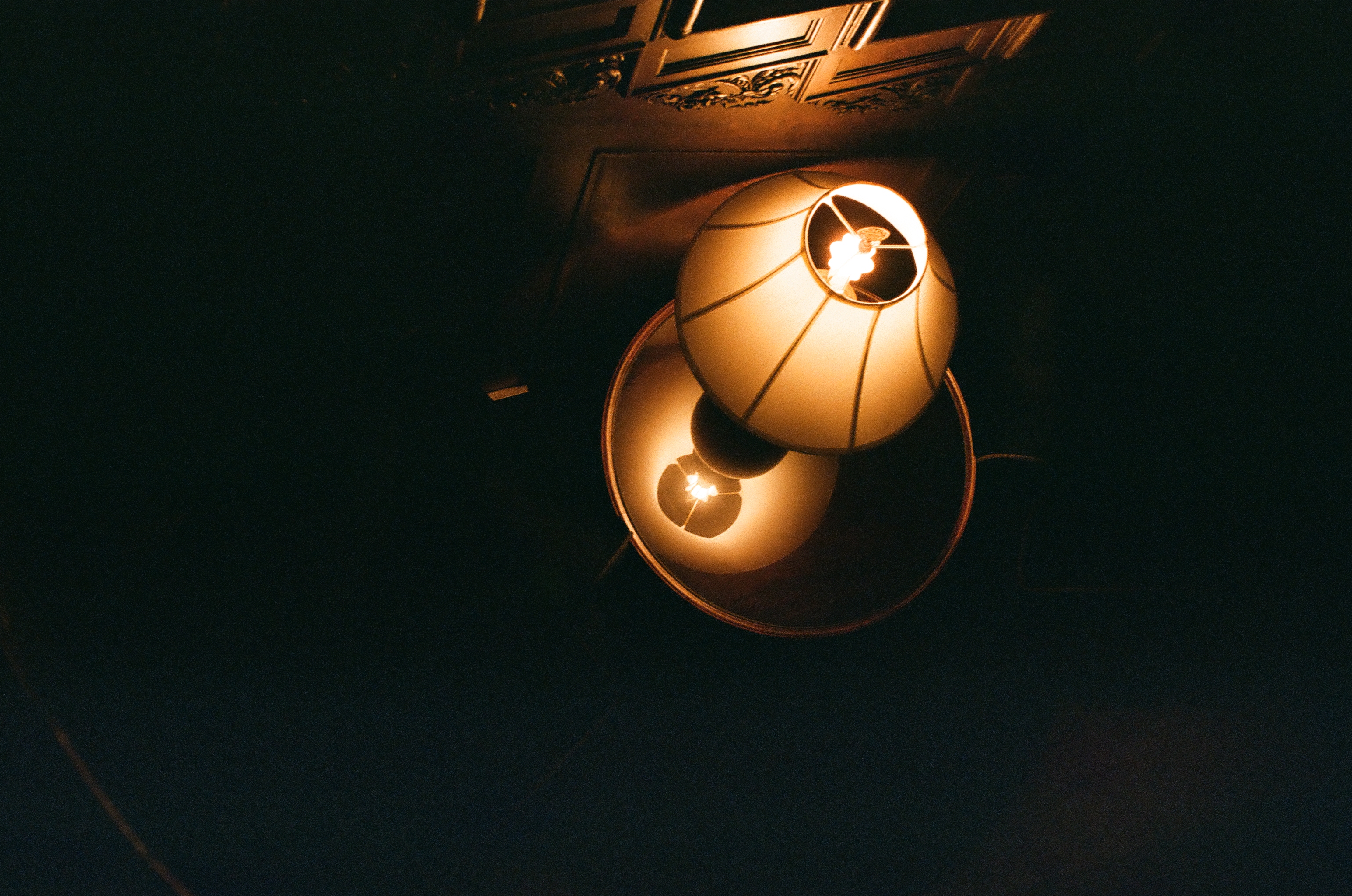 Two globe lamp shades glow warmly, one stacked above its own mirrored reflection, beneath dark carved ceiling molding.