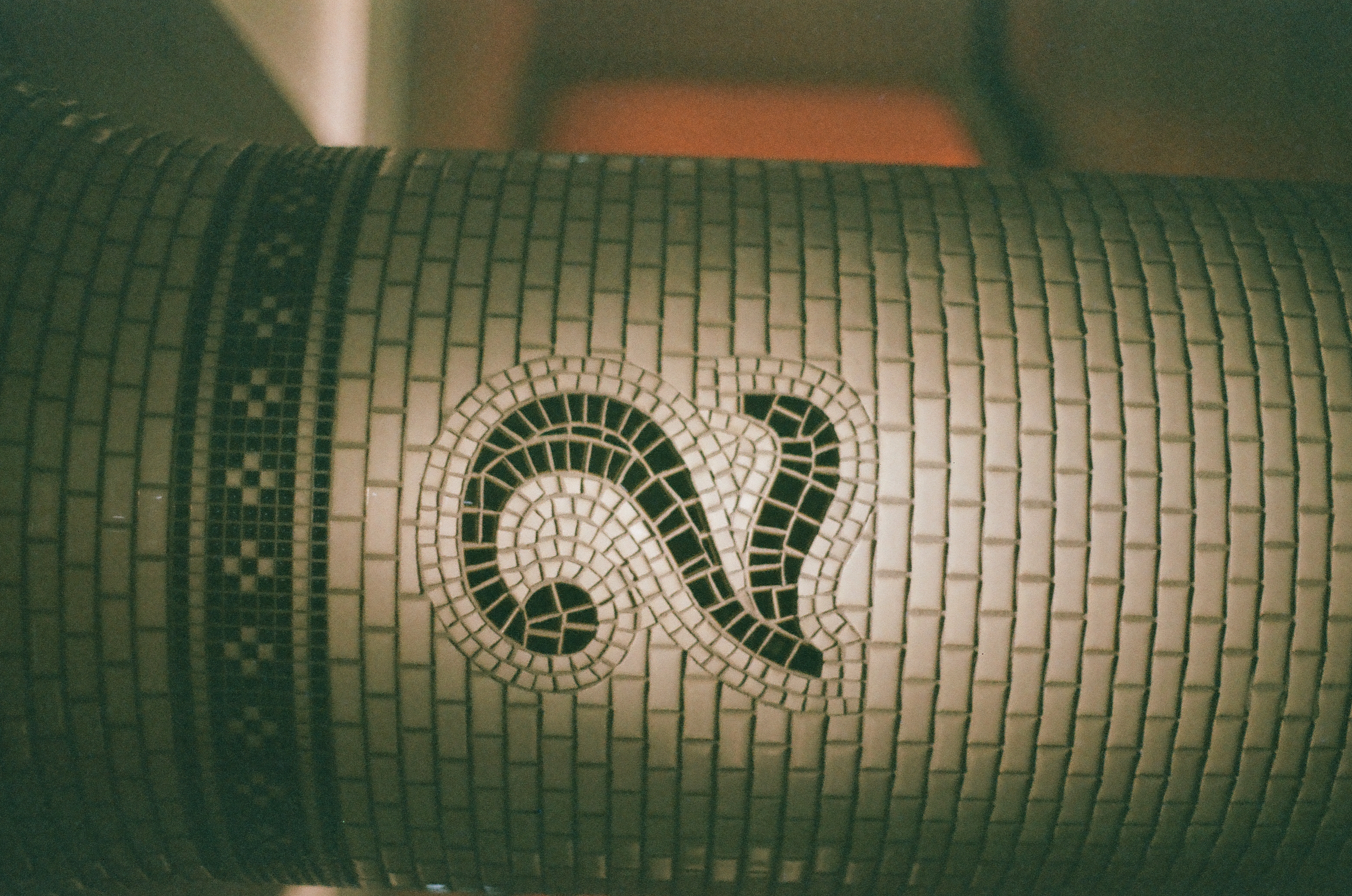 A tiled column shows the numeral two formed from dark mosaic tesserae in a curved shape beneath a checkered band, with an orange-lit area blurred behind it.