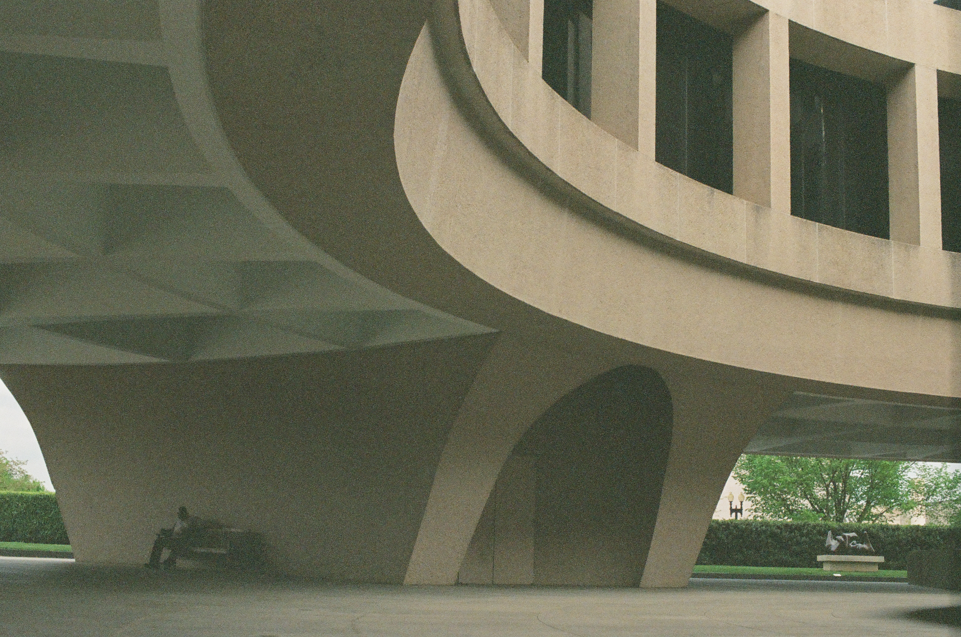 A broad circular concrete building rests on angled supports above benches and a small seated figure.