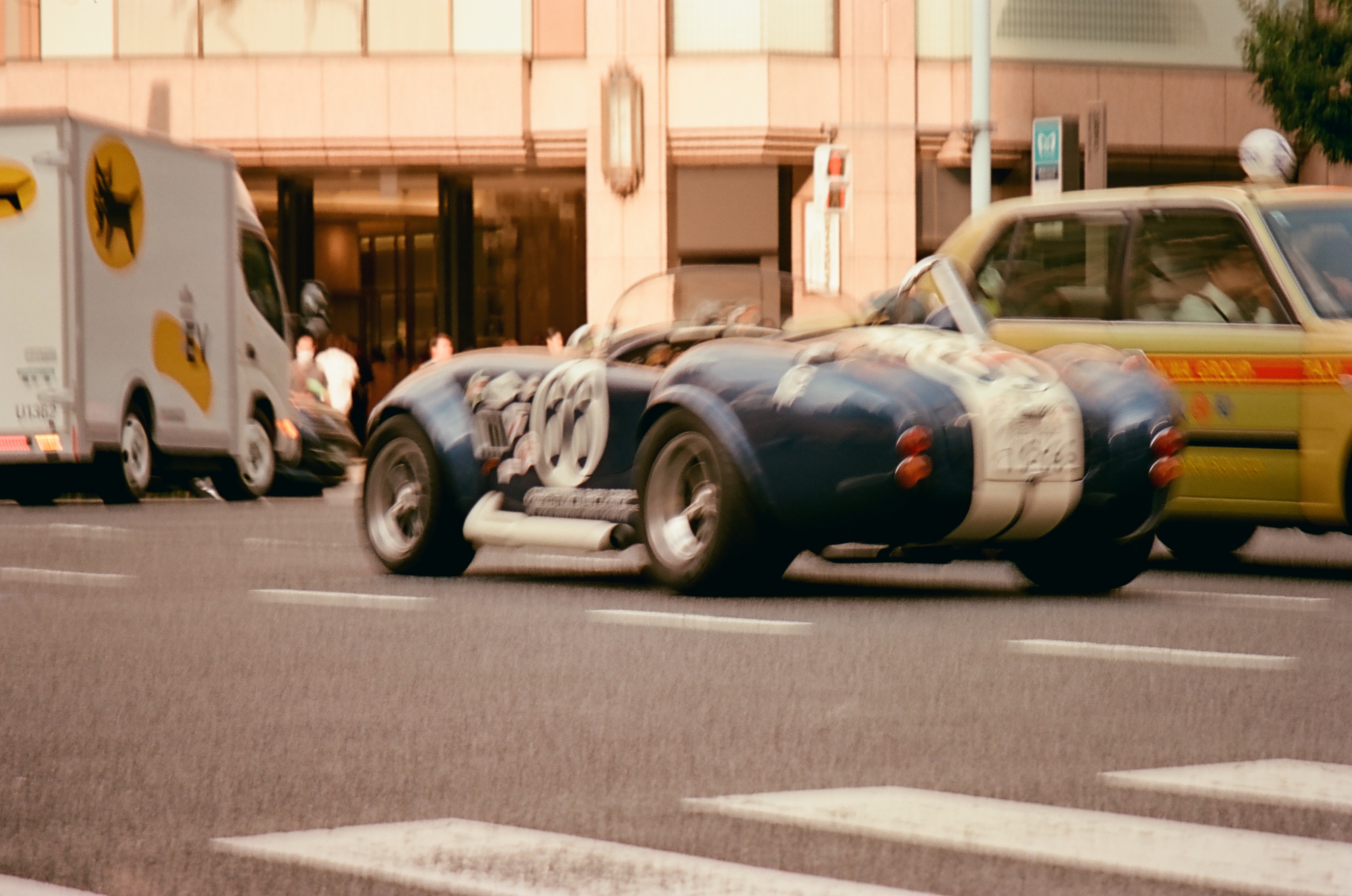 A dark blue open-top sports car with white stripes crosses a striped crosswalk between a yellow sedan with a roof sign and a white truck bearing a black cat emblem.