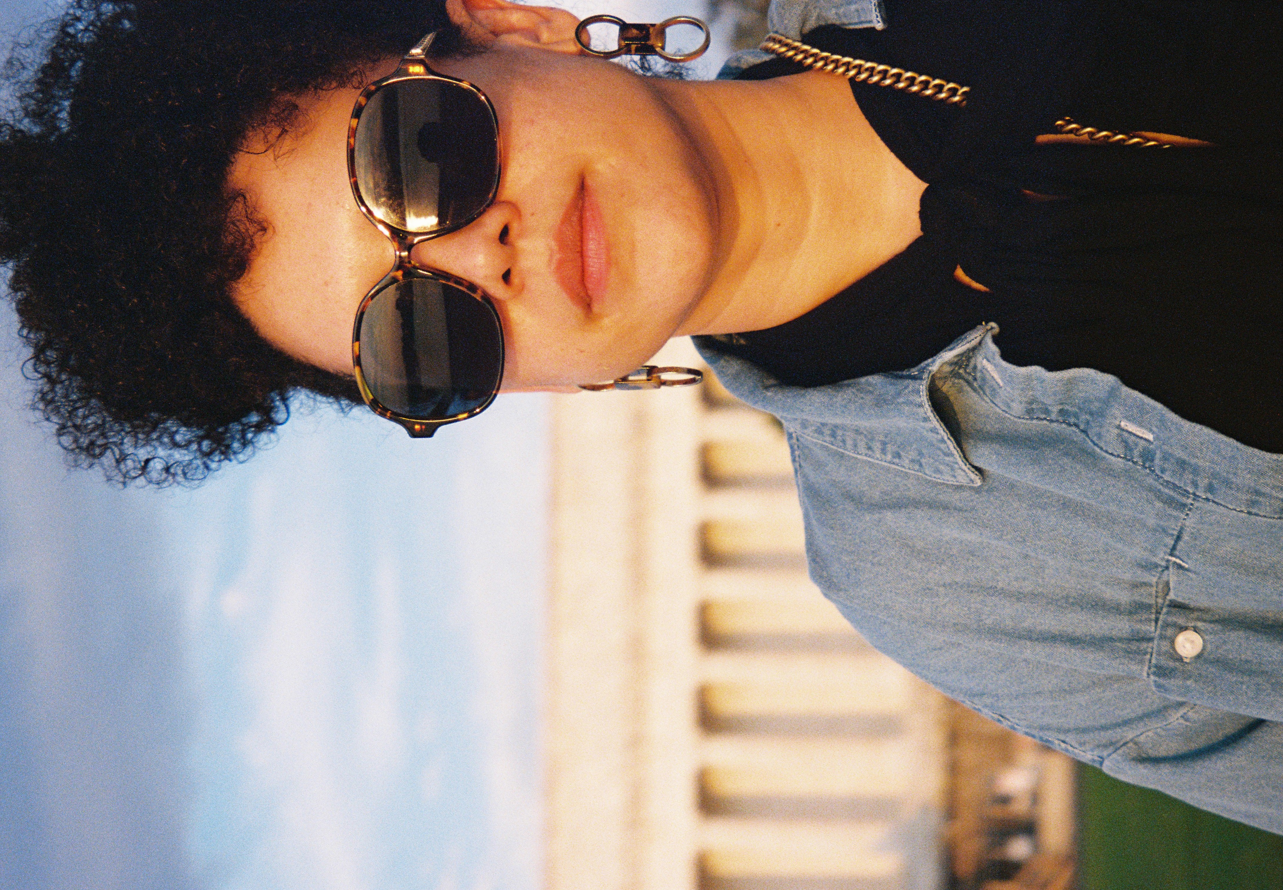 A woman in large tortoiseshell sunglasses, hoop earrings, a chain necklace, and a denim jacket faces the camera before a blurred columned building.