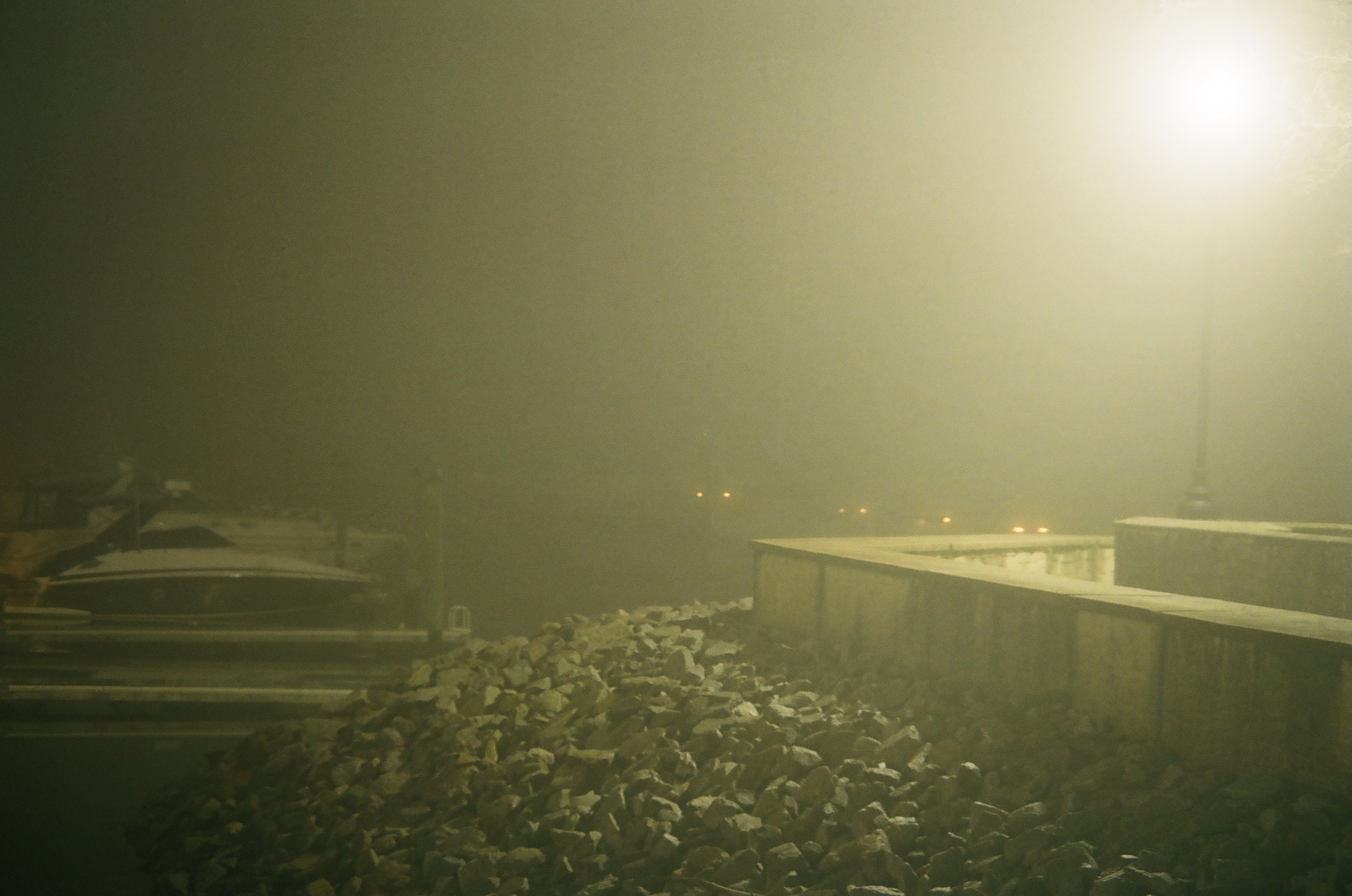 A bright streetlight glows through thick fog above a rocky shoreline, with a moored boat and dock visible at left.