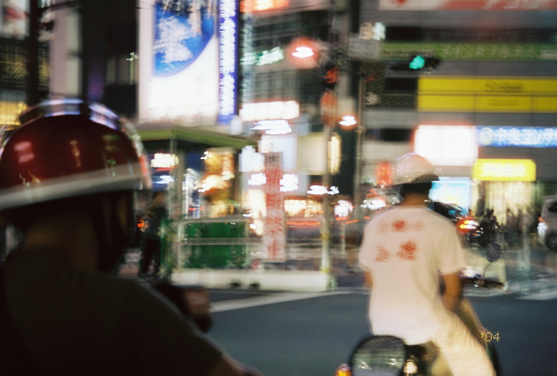 Seen past a red helmet in the foreground, another rider in a white t-shirt with red printed text waits at a night intersection.