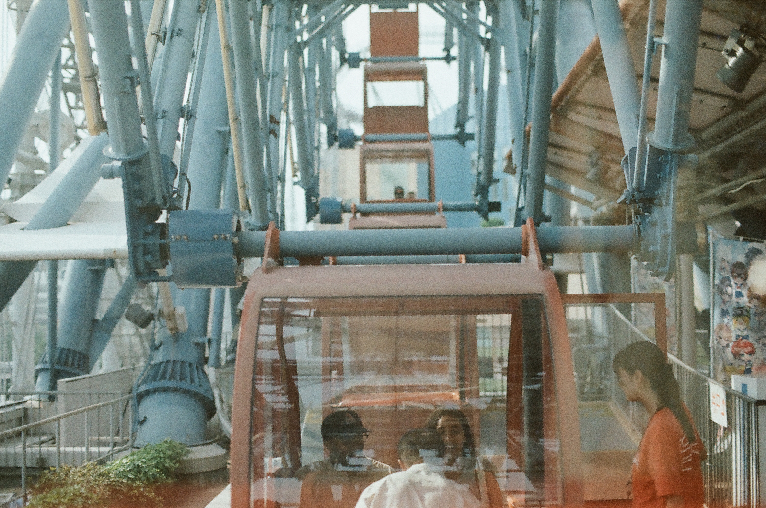 An orange gondola climbs past pale-blue steel supports on a Ferris wheel, with riders visible through its glass and a woman waiting at the platform.