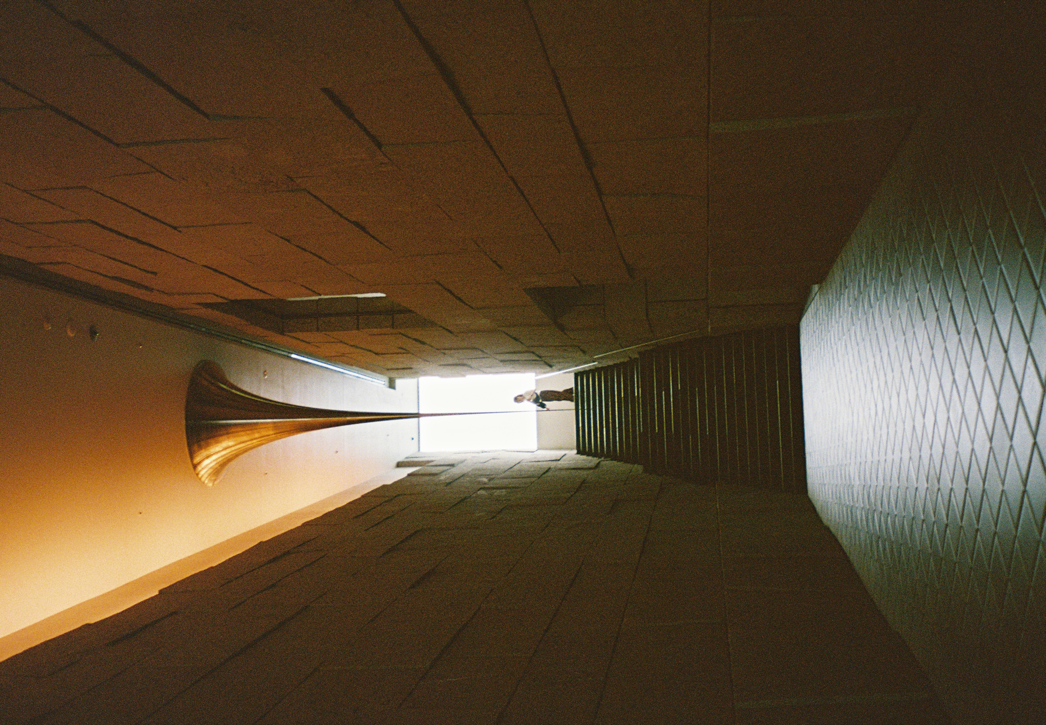 A narrow stone-walled stairwell climbs toward a bright doorway where one small figure stands, lit by a golden funnel-shaped ceiling fixture.