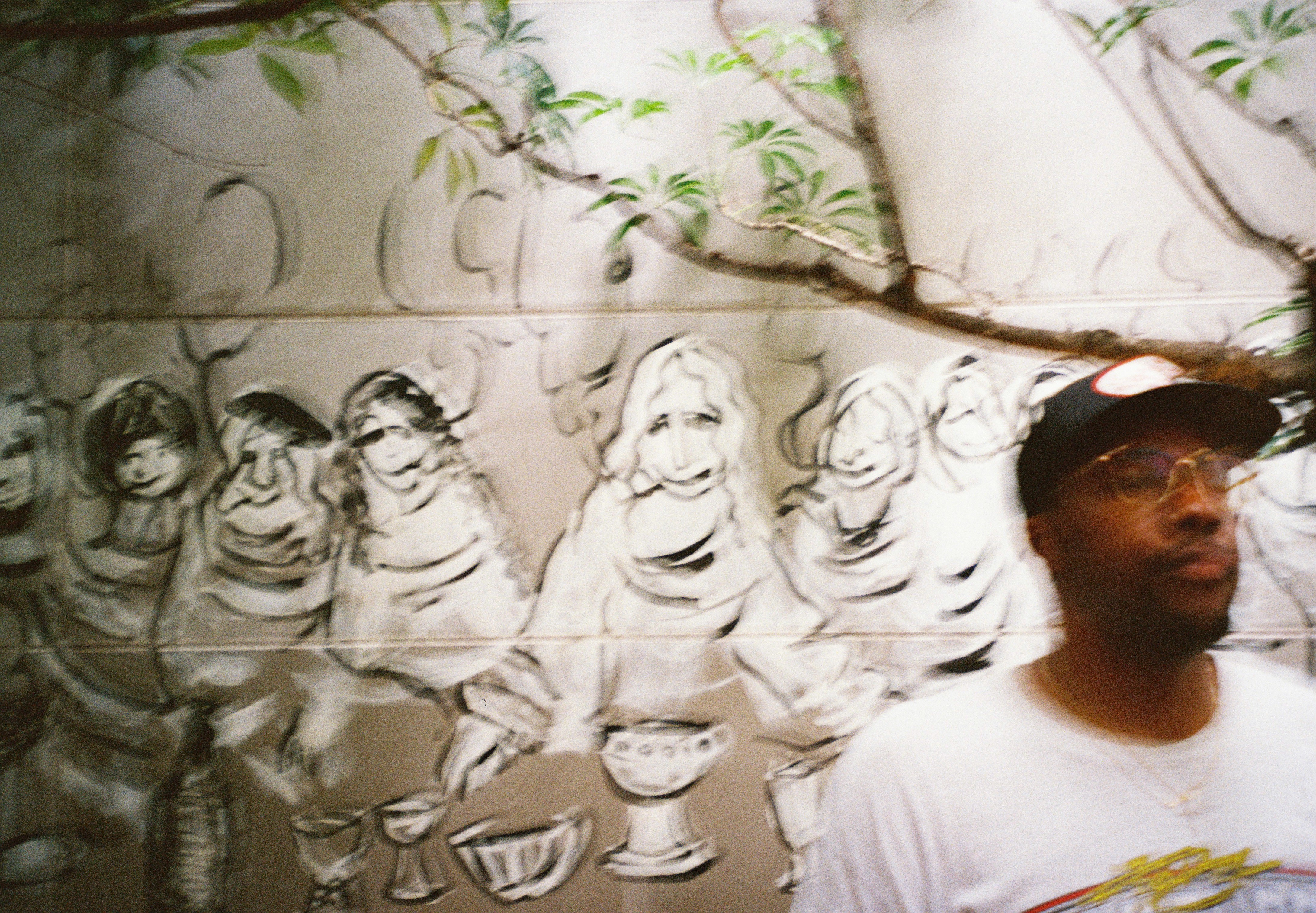 A man in a cap and glasses passes in front of a wall painted with loosely sketched robed figures and drawn drinking vessels.