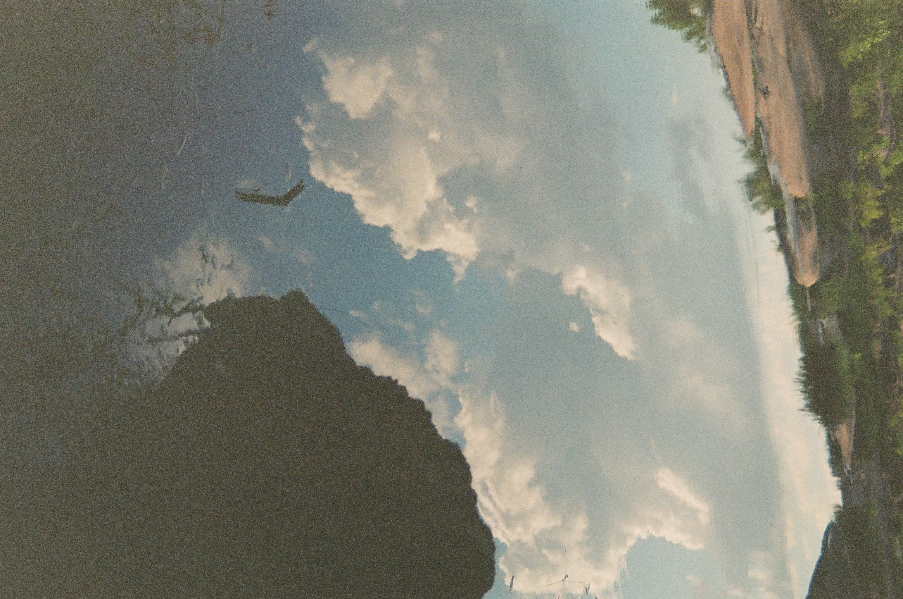 A blue sky and large white clouds reflect in still water beneath a rocky shoreline, with a dark boulder breaking the surface at right.