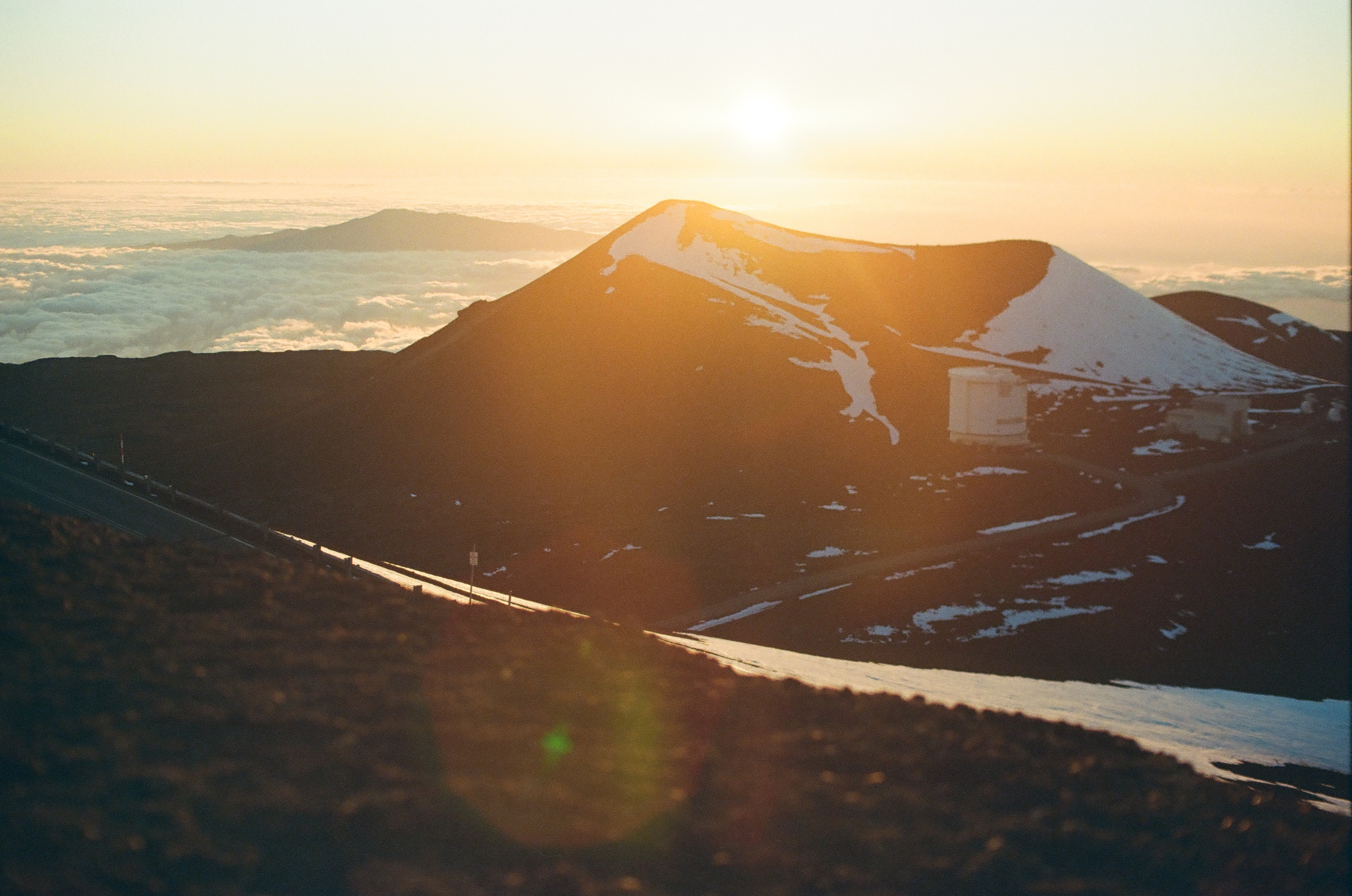 Low sun flares above a snow-lined ridge while a small pale structure sits in the shadowed basin.