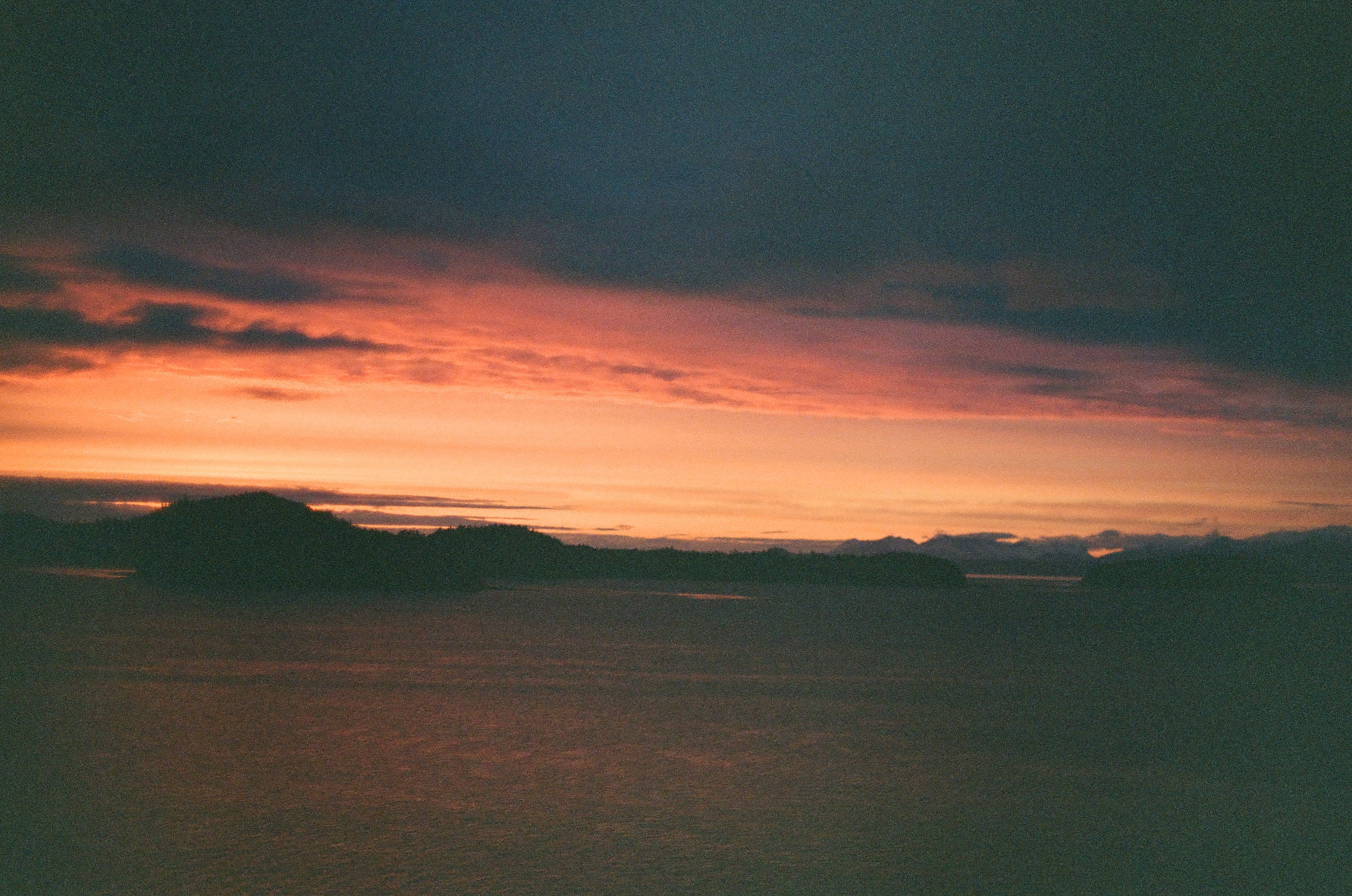 Heavy blue-gray cloud hangs over a saturated orange horizon, dark islands, and gently rippled water.
