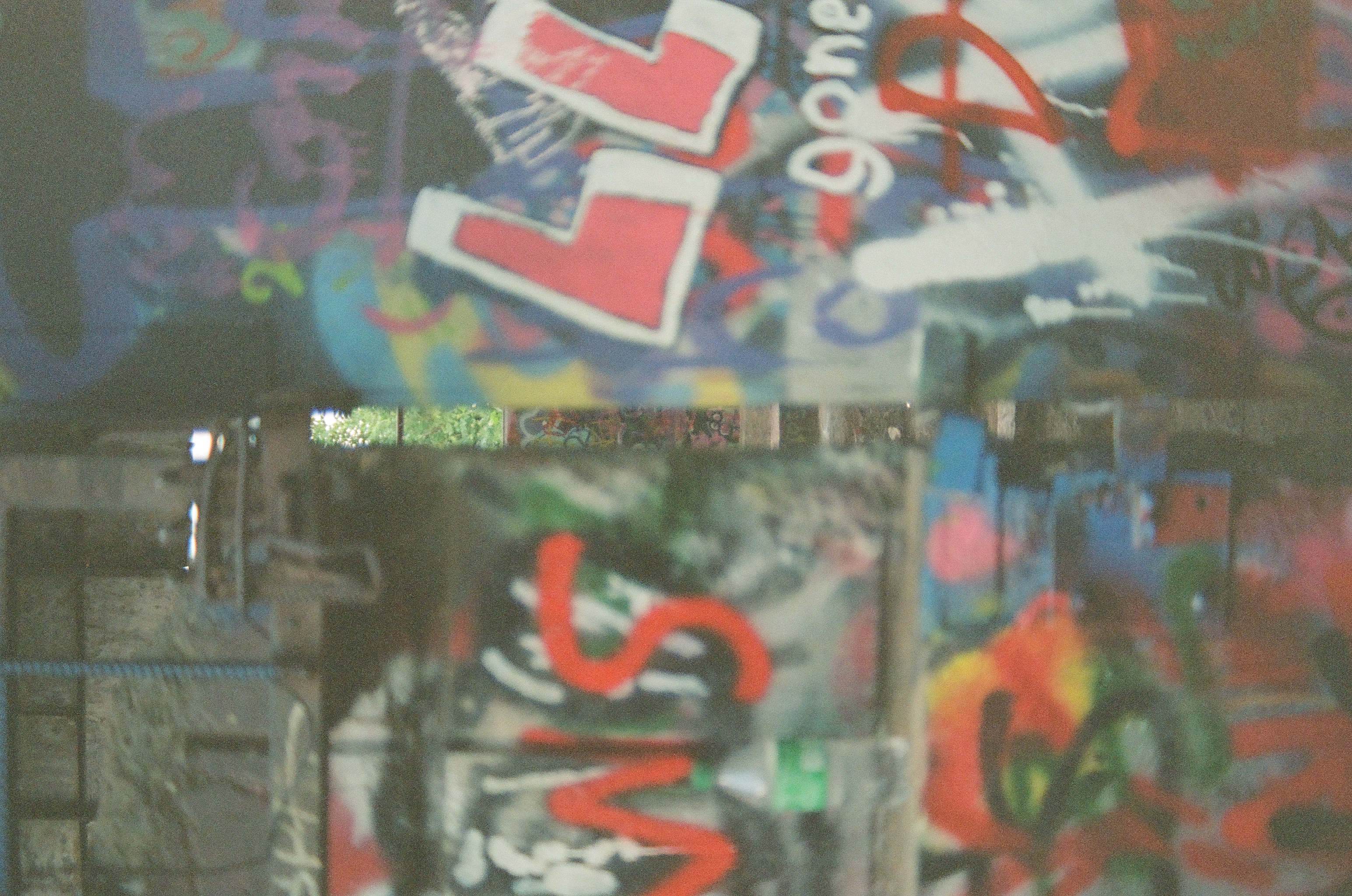 Fragments of red and white letters overlap blue, green, and black painted marks behind reflective glass.